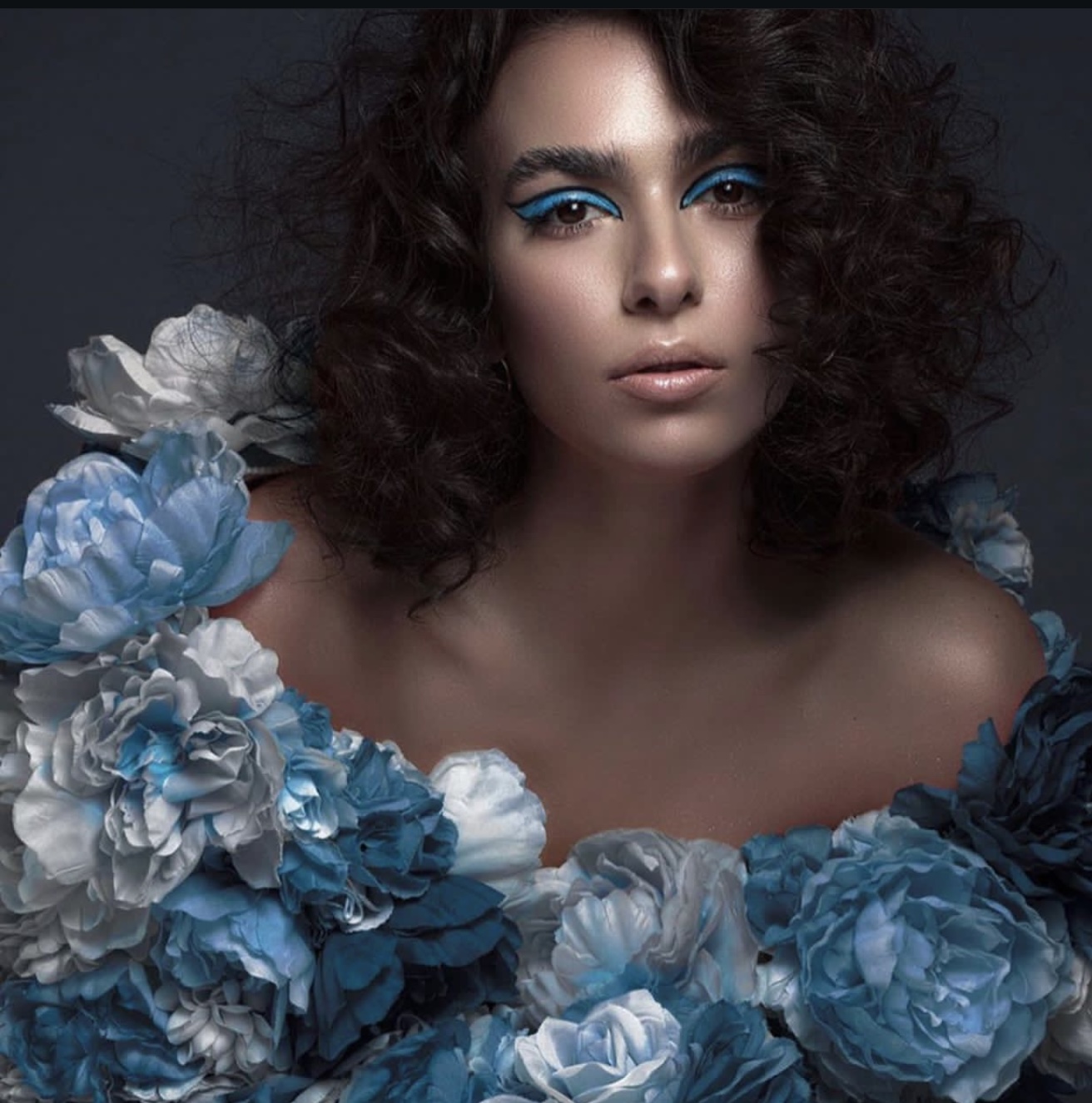 Woman with curly hair and blue makeup surrounded by blue and white flowers, looking at the camera.