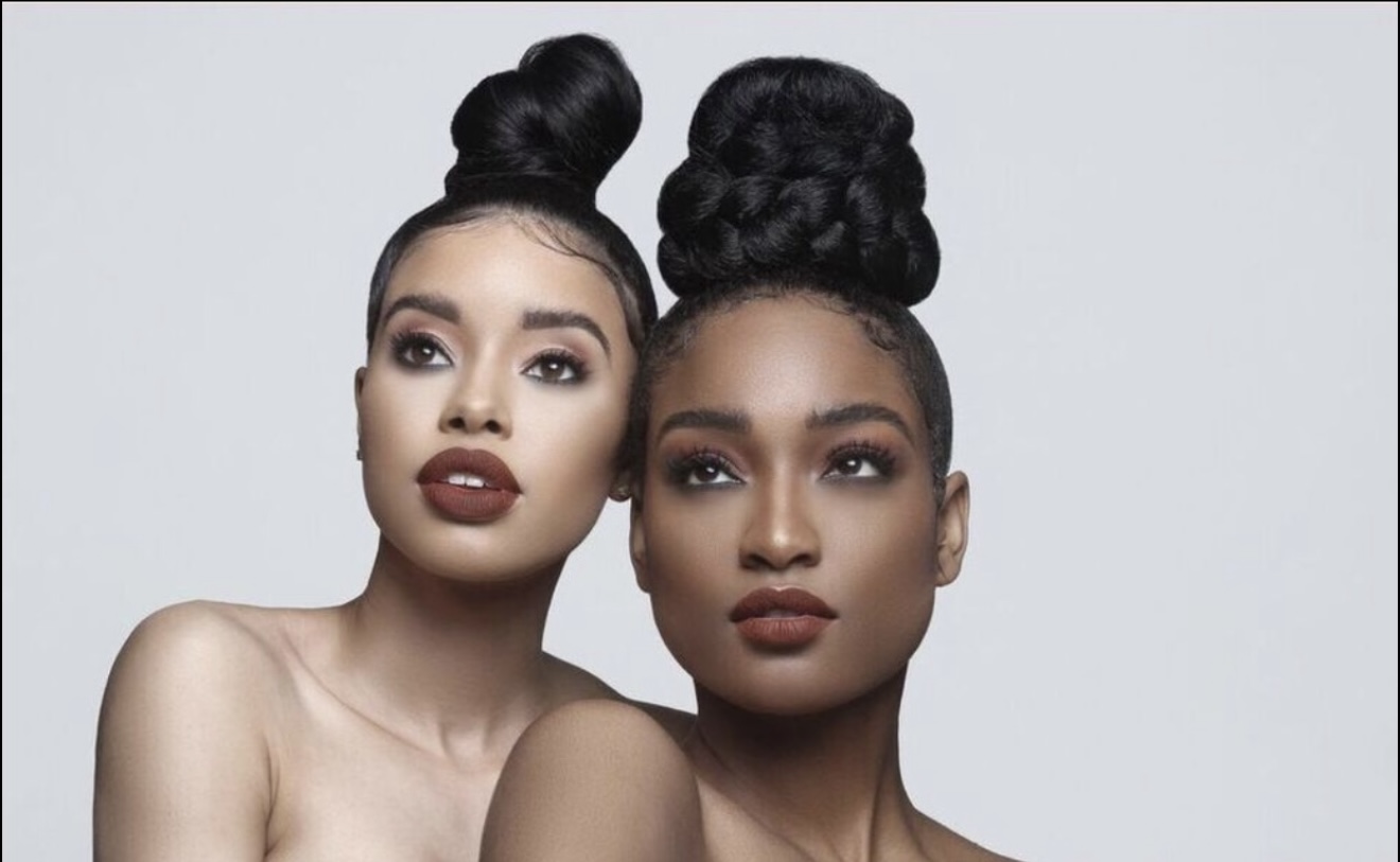 Two women with styled hair and makeup pose closely against a plain background.