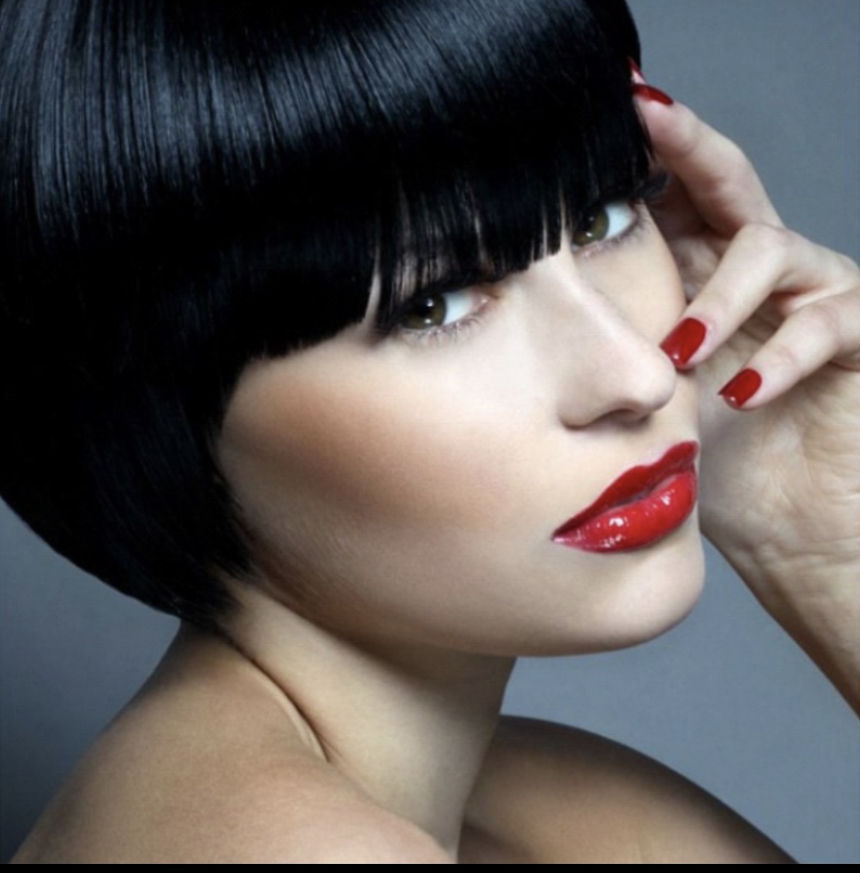 Woman with short black hair, red lipstick, and red nail polish, touching her face with her hand.