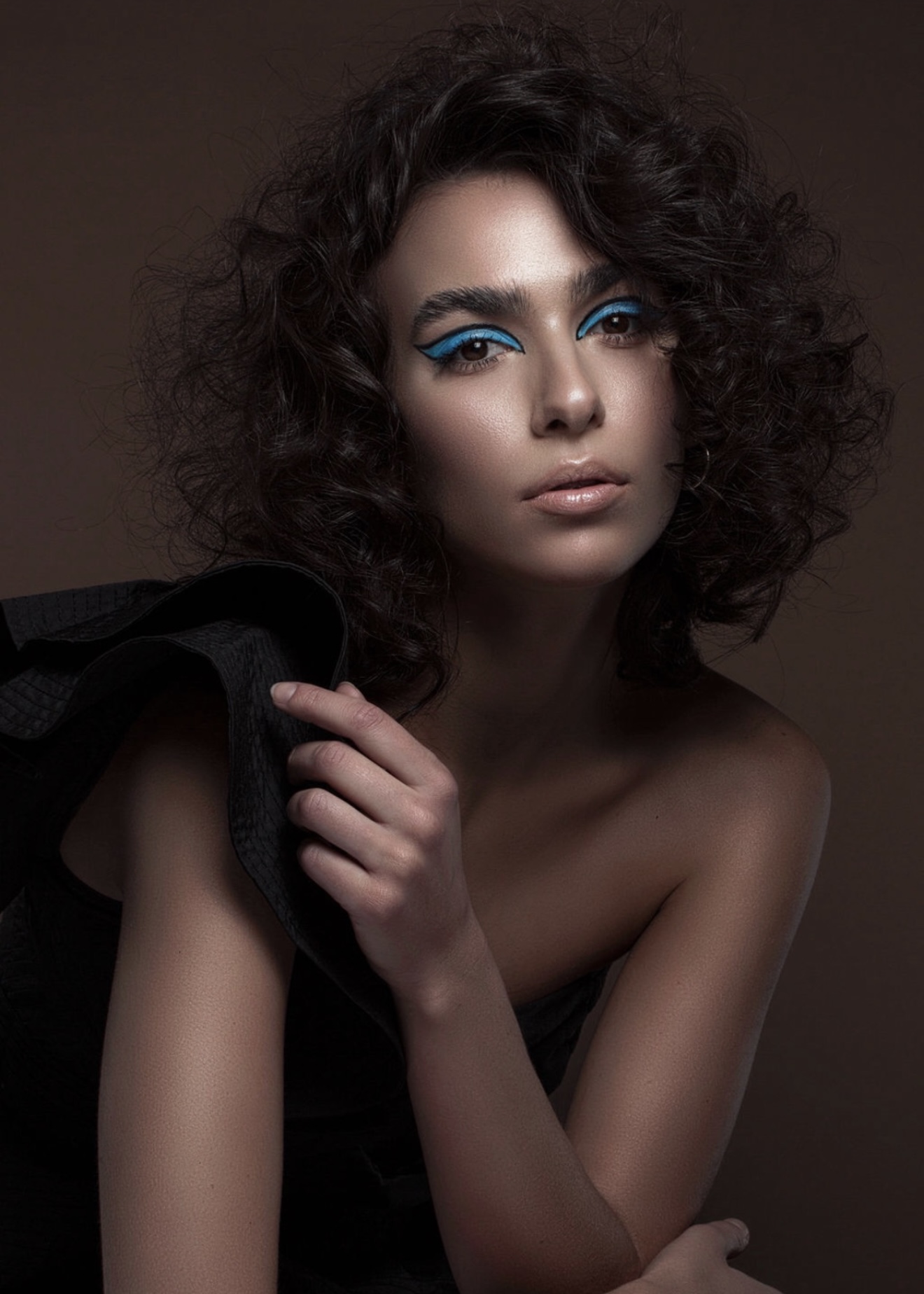 Young woman with curly hair and blue eye makeup, looking at the camera, with a dark background.