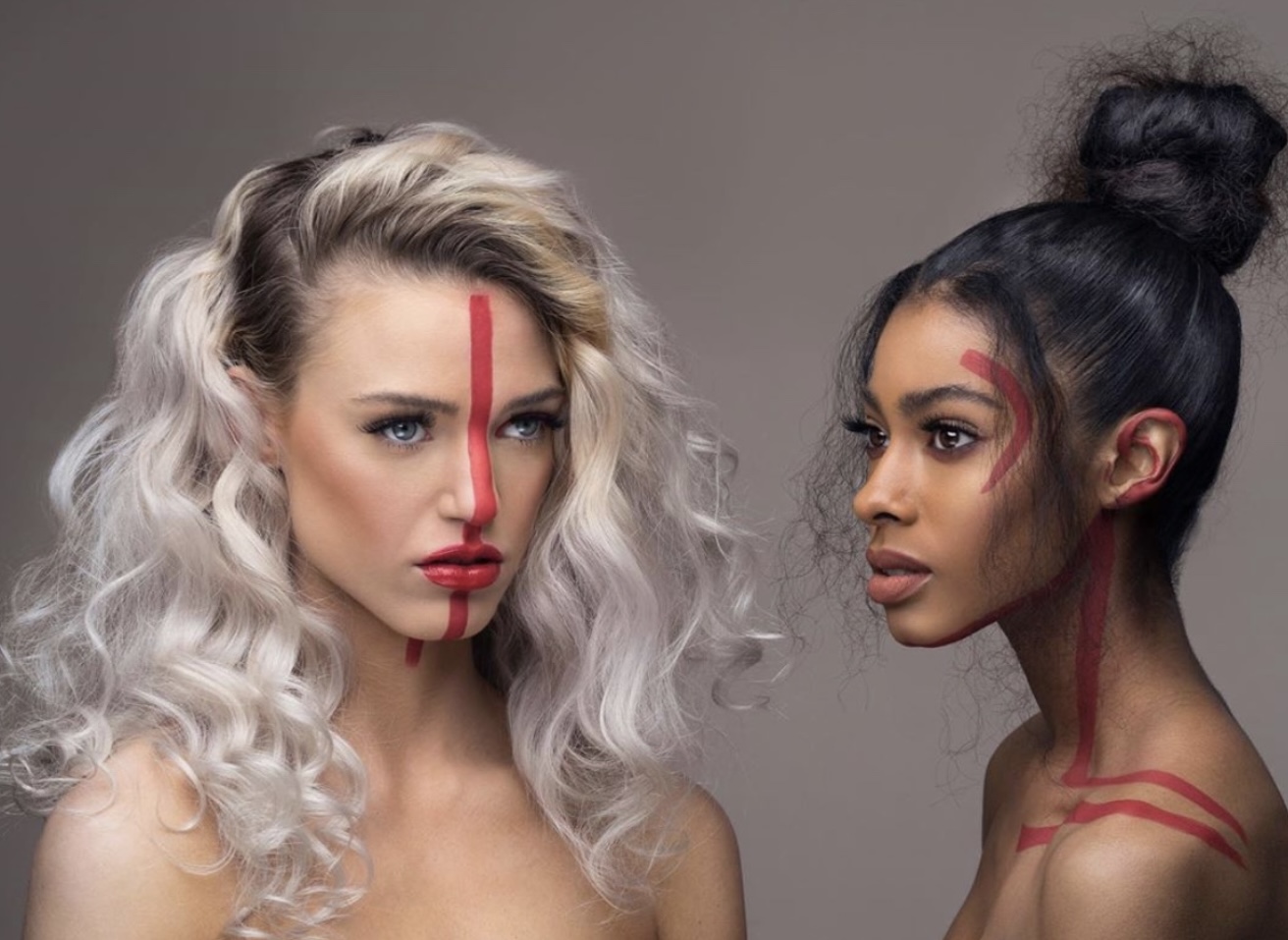 Two women with artistic red face and body paint, one with curly blonde hair and the other with dark hair in a bun, face each other.
