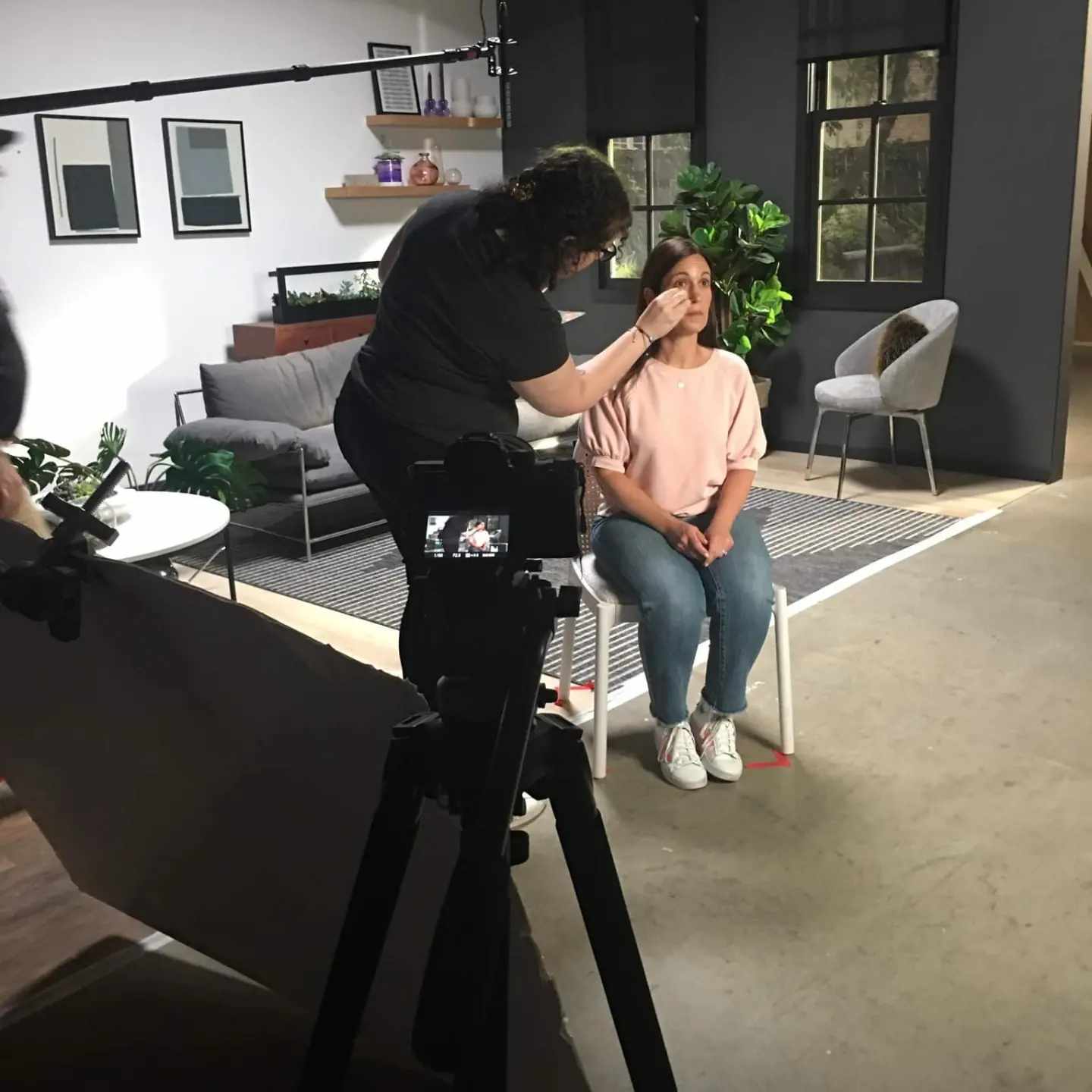 Makeup artist applying makeup to a seated woman in a studio setting with furniture and plants.