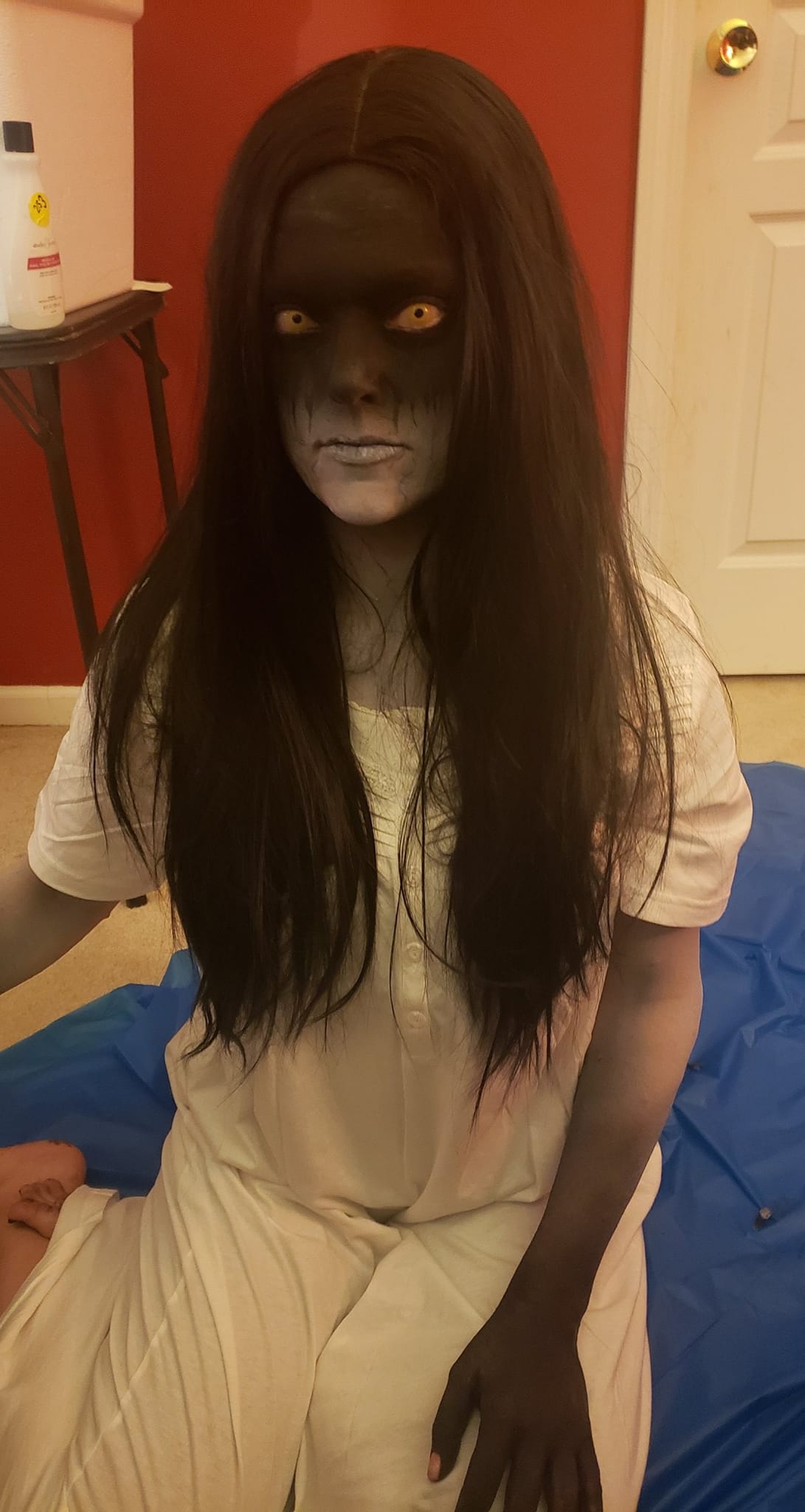 Person with long dark hair and face painted to resemble a skull, sitting indoors on a blue mat, wearing a white outfit.