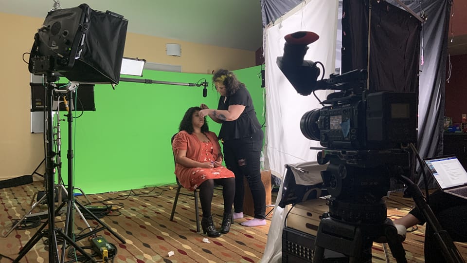 A woman is applying makeup to a seated woman in front of a green screen, with filming equipment around them.