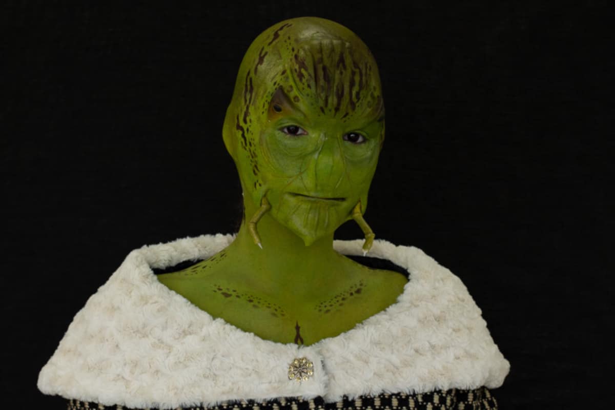 A person dressed as a green alien with a textured face, wearing a white fluffy garment, against a black background.