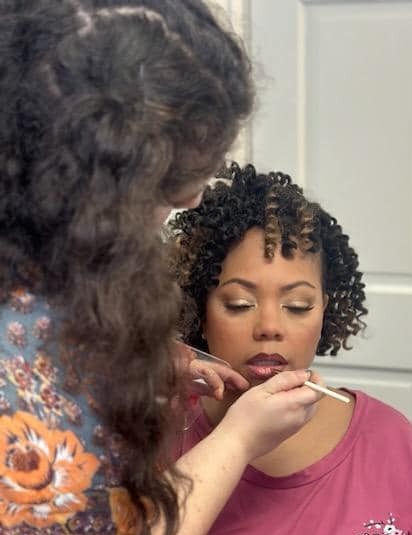 A makeup artist applies makeup to a woman with short curly hair, focusing on her face, in a close-up shot.