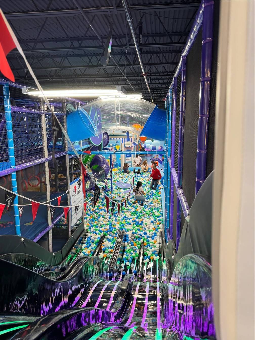 Indoor playground with colorful ball pit, slides, and children playing, viewed from top of a slide, vibrant and lively scene.