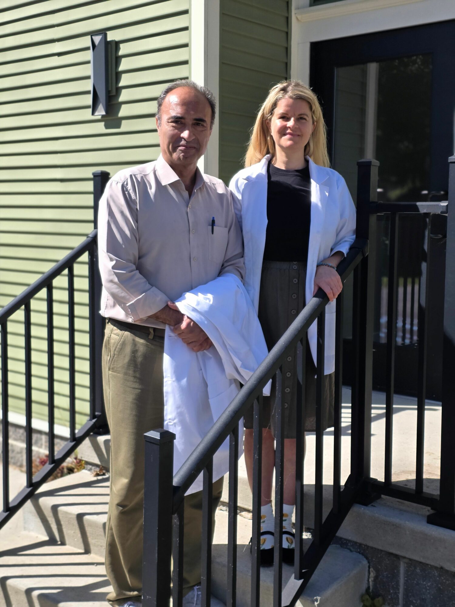 Two women standing on a porch, one in a white coat and the other in a white dress, with a black railing.