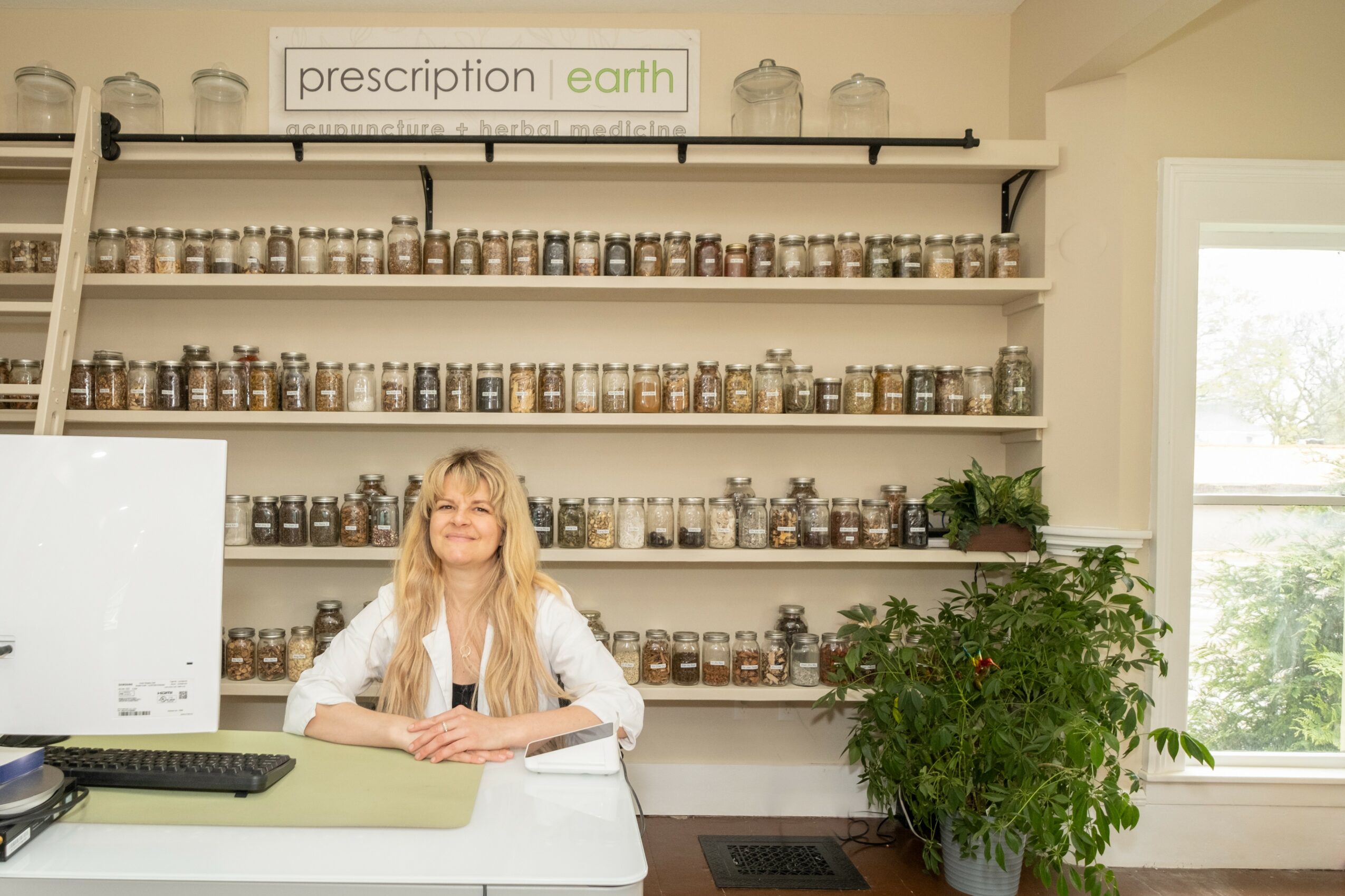 Woman with long blonde hair sitting at a counter in front of shelves filled with jars, next to a large green plant, window on right