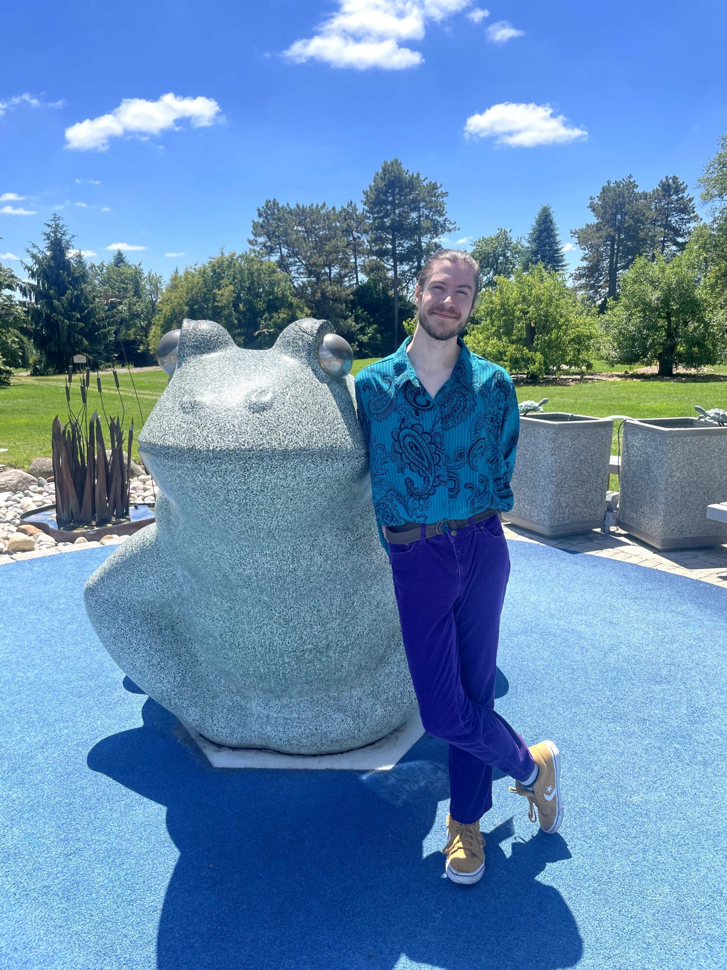 Person standing next to a large frog sculpture outdoors on a sunny day with trees and blue sky.