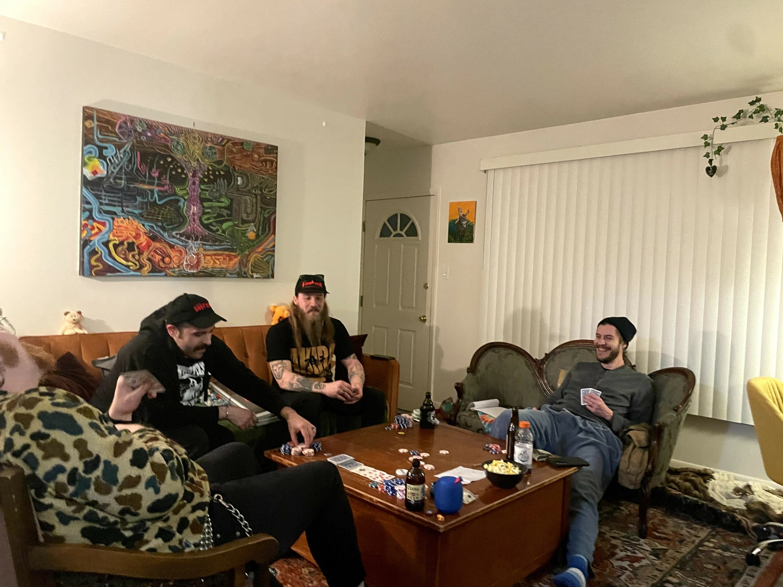 Four people sitting in a living room, two on a sofa, one on an armchair, and one on a chair, with a coffee table in front, a colorful painting on the wall, and a window with vertical blinds.