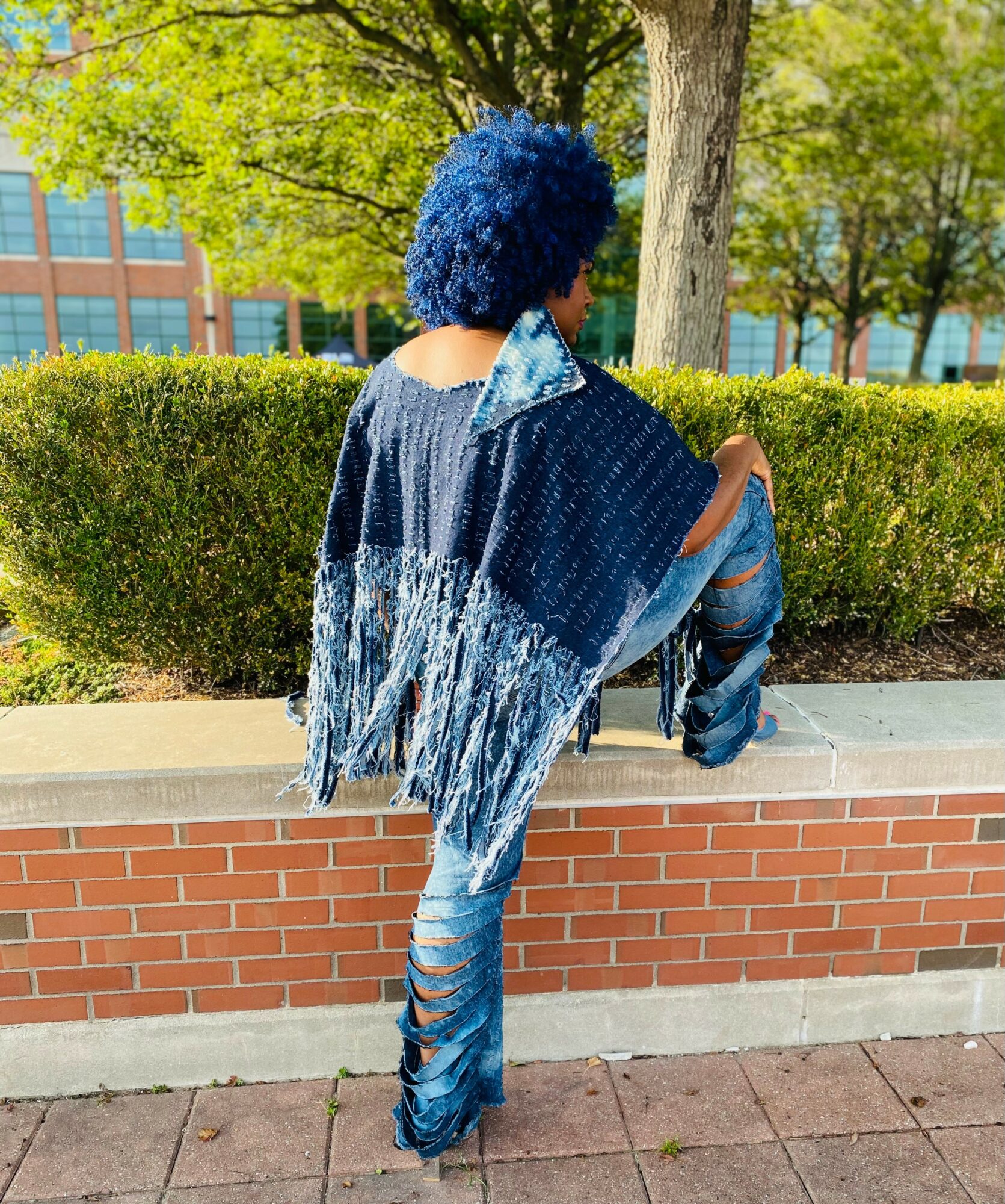 Person with blue curly hair wearing a denim jacket and ripped jeans, standing outdoors near a brick wall and greenery.