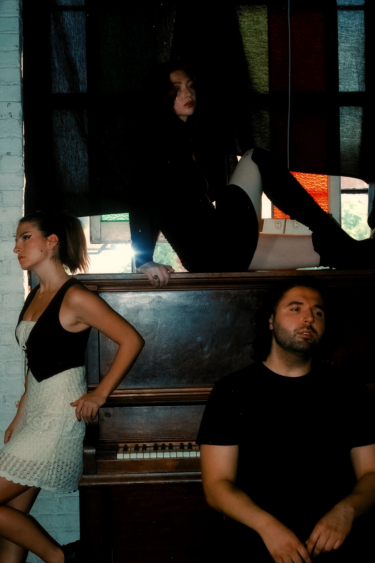 Three people near a piano, one sitting, one standing, and one on a ledge, in a dimly lit room.