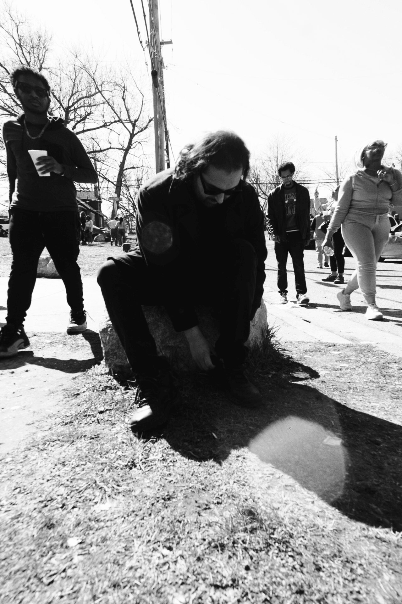 Person planting a small tree or shrub on the ground outdoors, with several people nearby, in a black-and-white photo.