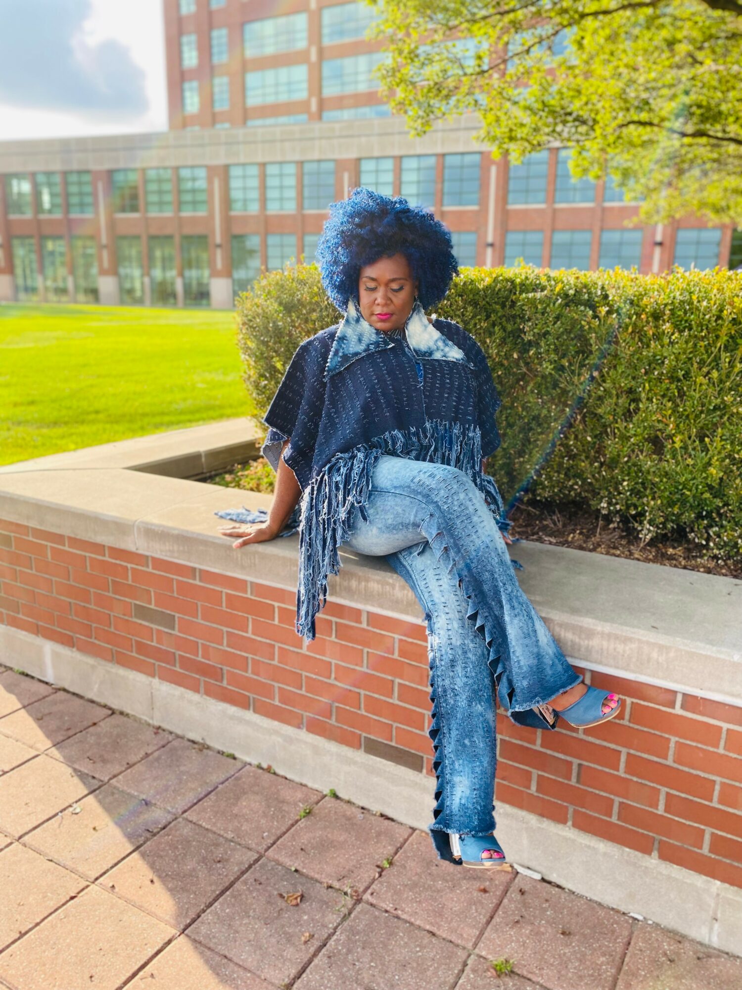 Woman with blue curly hair sitting on a brick ledge outdoors, wearing a dark top and jeans, with greenery and a building in background.