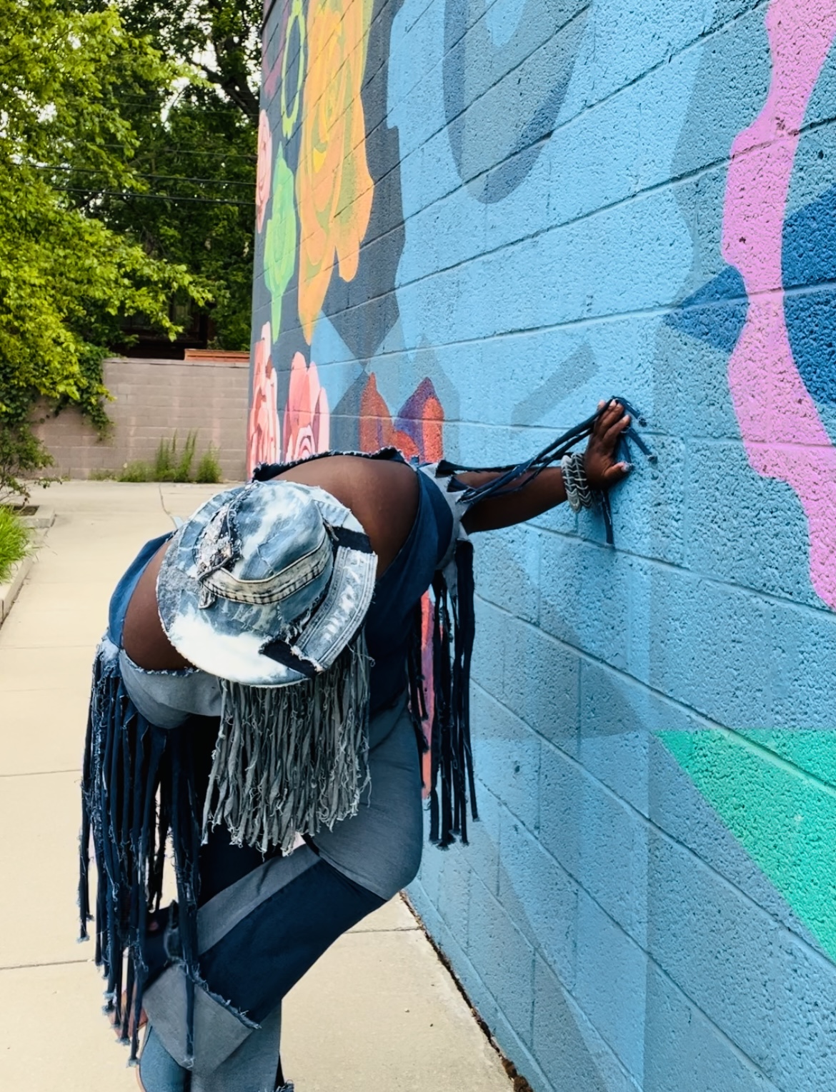 Person painting a colorful mural on a blue wall, wearing a hat and dark clothing, leaning forward with one hand on the wall.