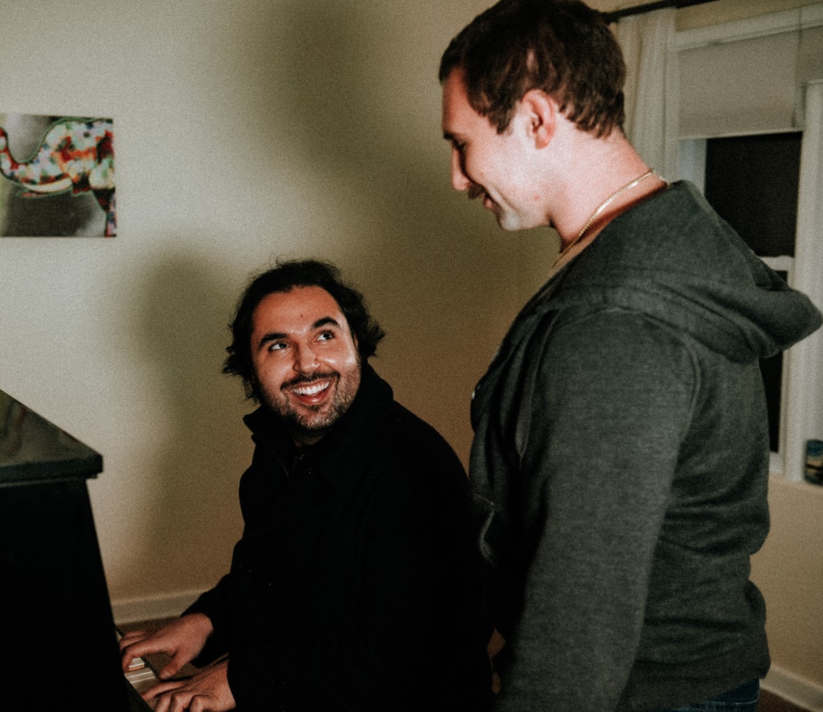 Two men interact indoors, one seated at a piano and the other standing, smiling at each other.