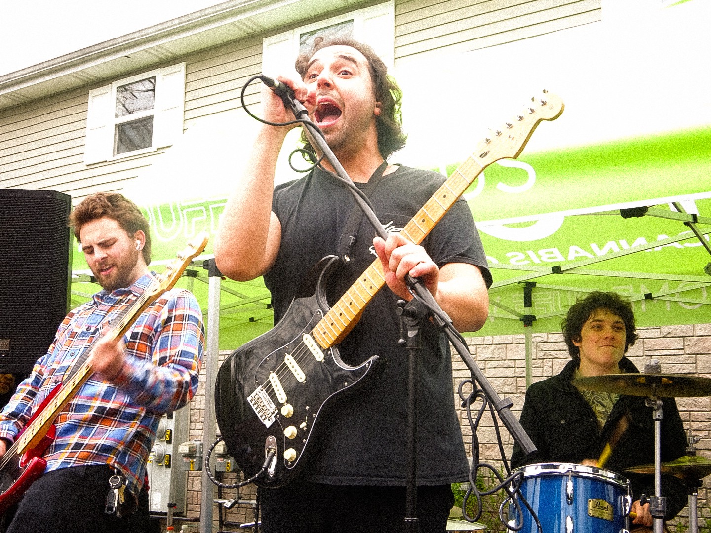 Three musicians perform outdoors, one singing into a microphone, another playing guitar, and a third on drums, under a green canopy.