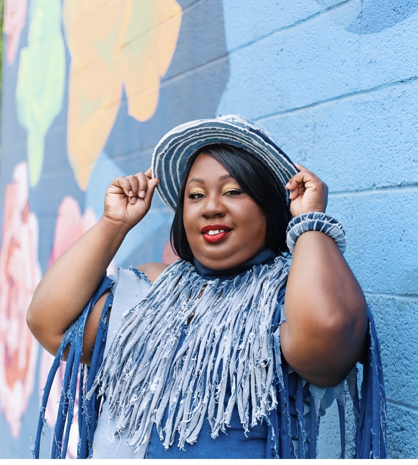 Woman wearing a striped hat and fringed dress, standing against a colorful mural wall, smiling, holding her hat.