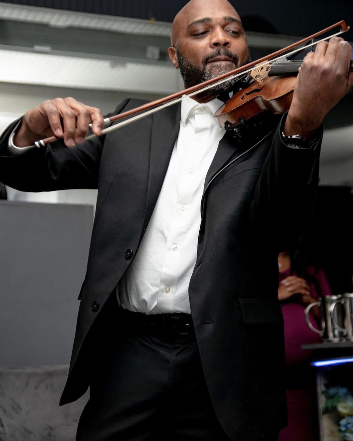 Man playing violin, wearing a black suit and white shirt, with eyes closed, in a performance setting.