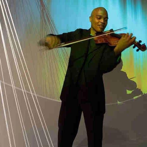 Person playing violin, standing with a colorful background, smiling, holding the violin and bow, wearing a dark suit.