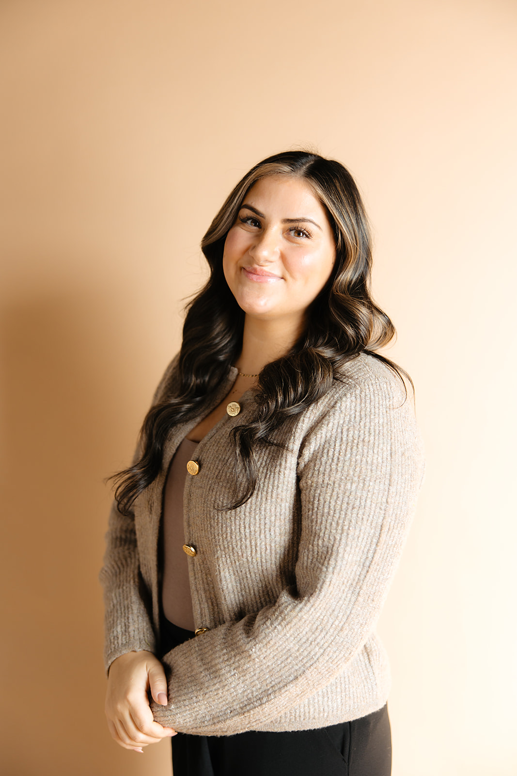 Young woman with long wavy hair smiling, wearing a beige cardigan with gold buttons, standing against a beige background.