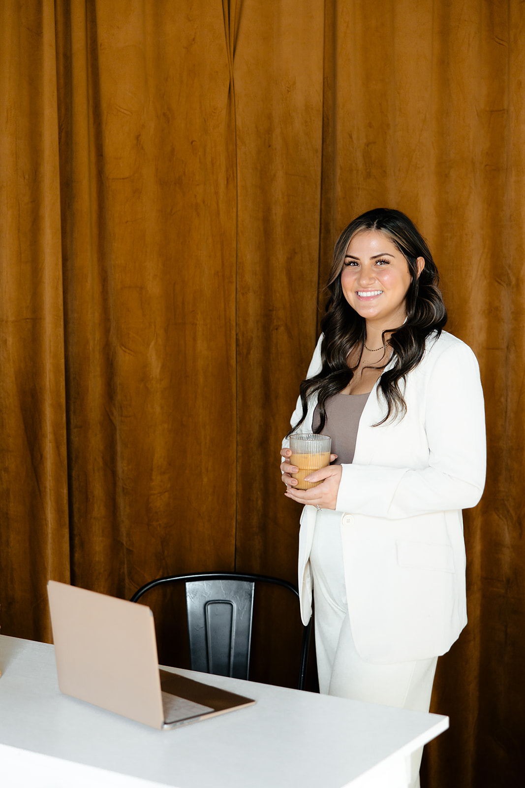 Smiling woman with long dark hair holding a drink, standing near a table with a laptop, against a wooden wall.