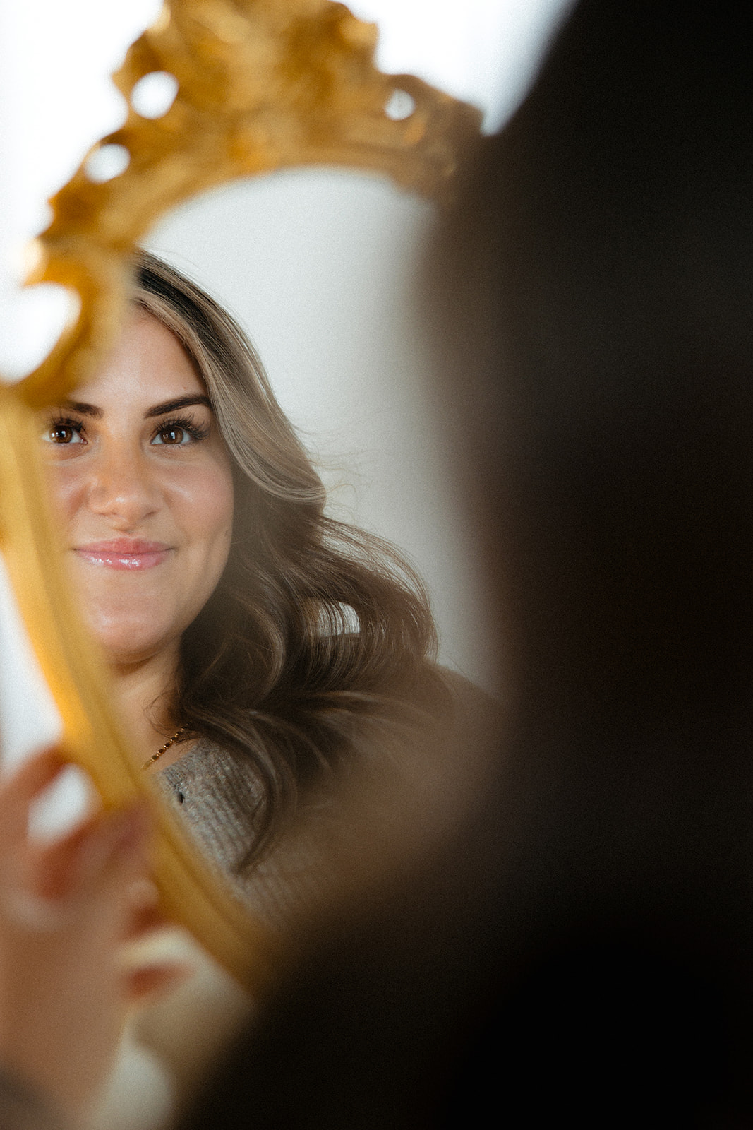 Woman with long wavy hair looking at her reflection in a mirror, smiling, with part of the mirror frame visible.