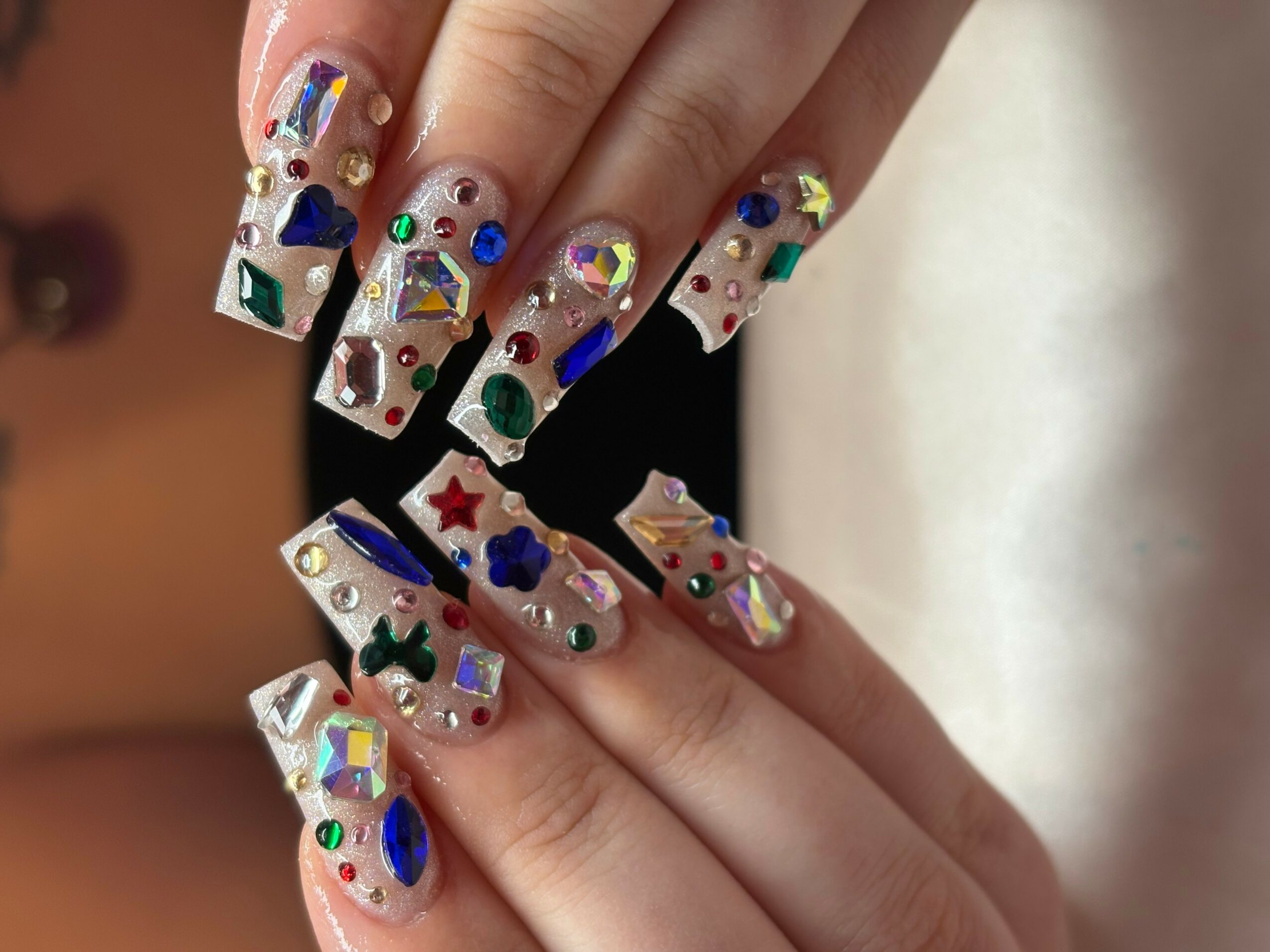 Hand with decorated nails featuring colorful shapes and rhinestones, held against a blurred background.