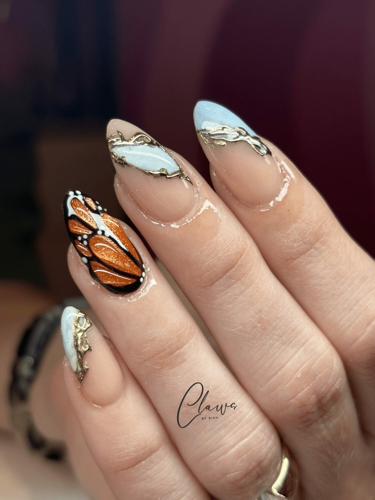 Close-up of a hand with butterfly and marble nail art, featuring orange, black, white, and gold colors, and a gold ring.
