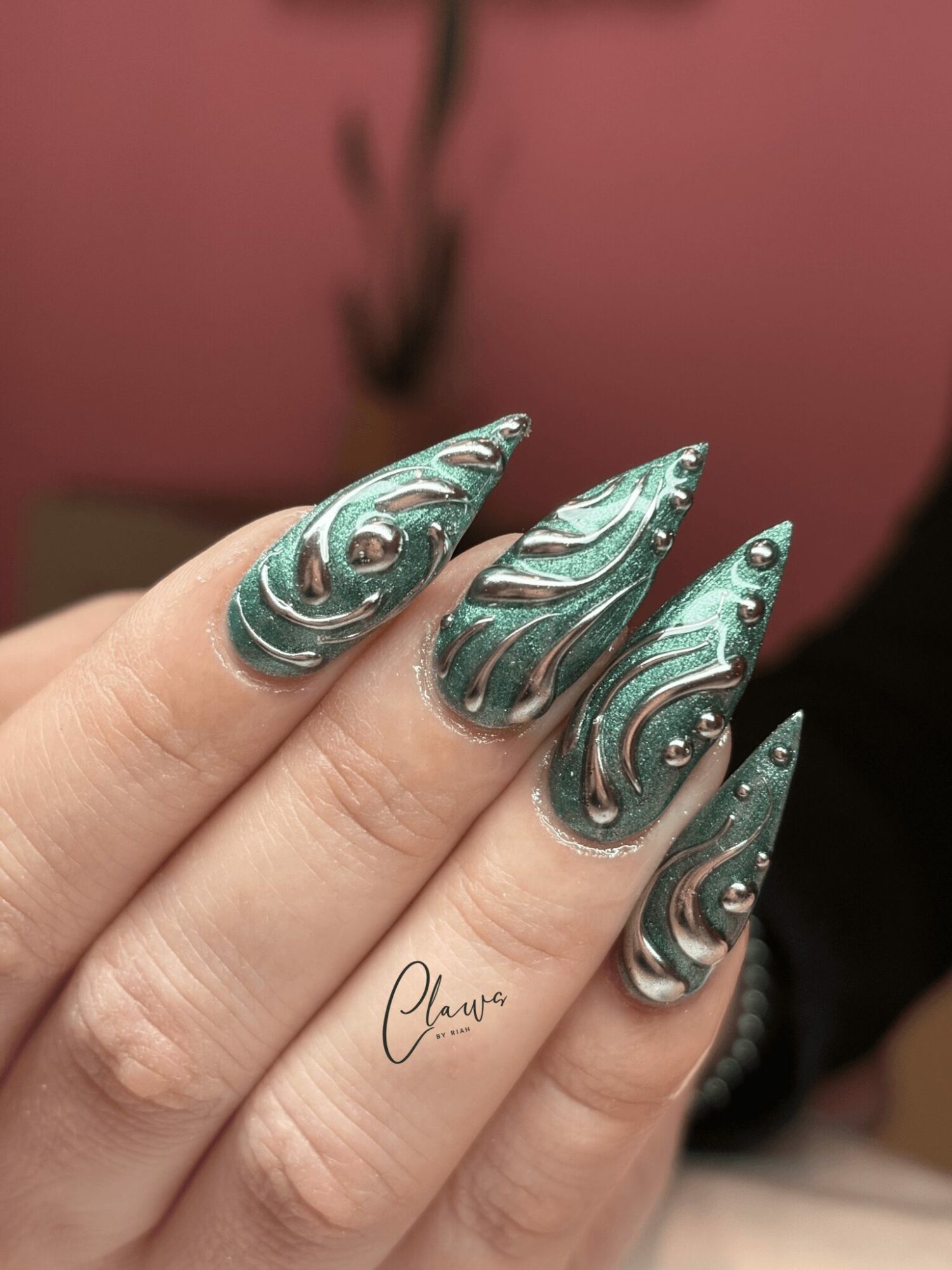 Close-up of five fingers with metallic, swirling nail art in teal and silver colors, against a pink background.