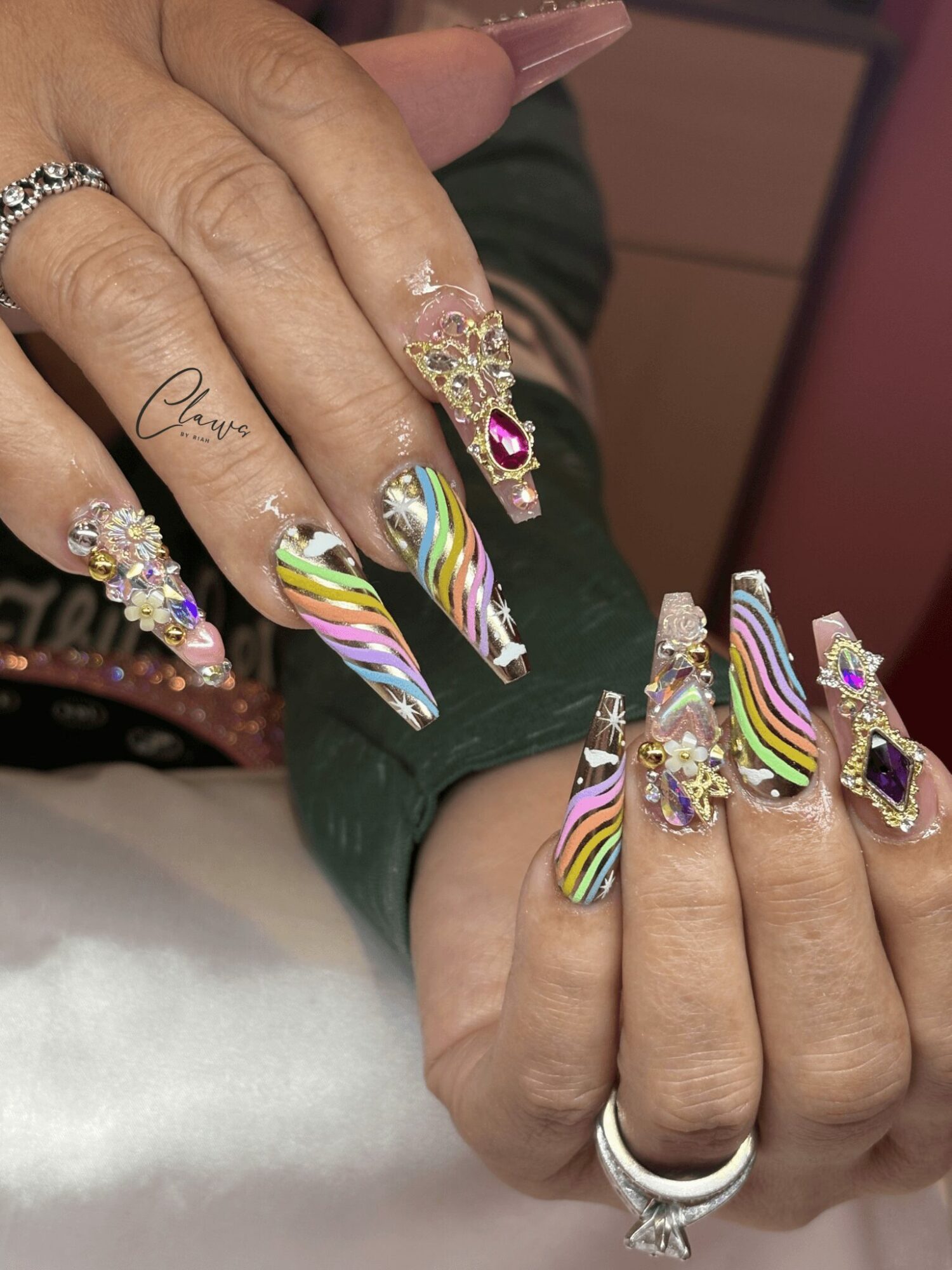 Close-up of hands with long, colorful, rainbow-striped nails and decorated rings, showing intricate nail art and jewelry.