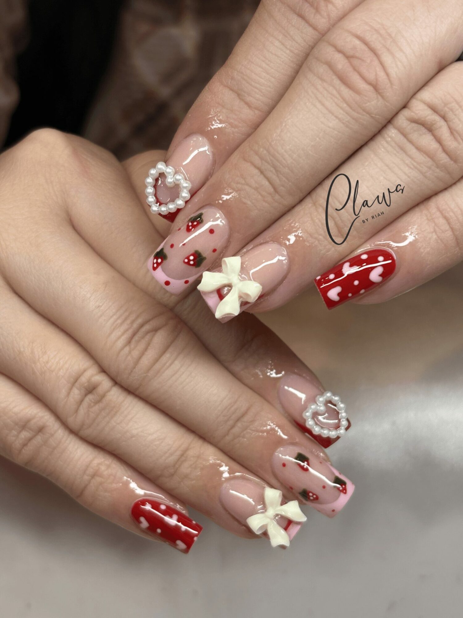 Close-up of hands with decorated nails, featuring red, white, and pink designs, bows, and heart-shaped accents.