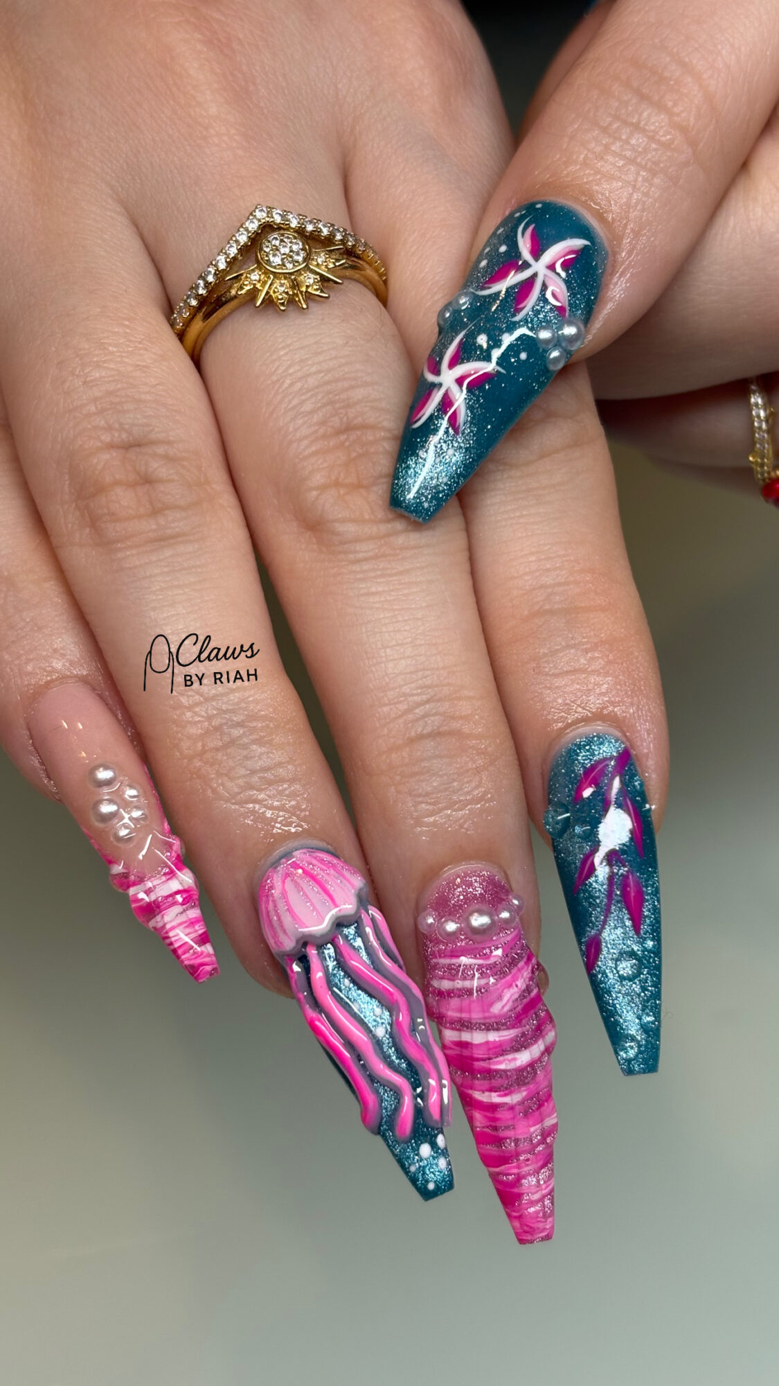 Close-up of hands with colorful, decorated long nails featuring pink, blue, and purple designs, and gold rings.
