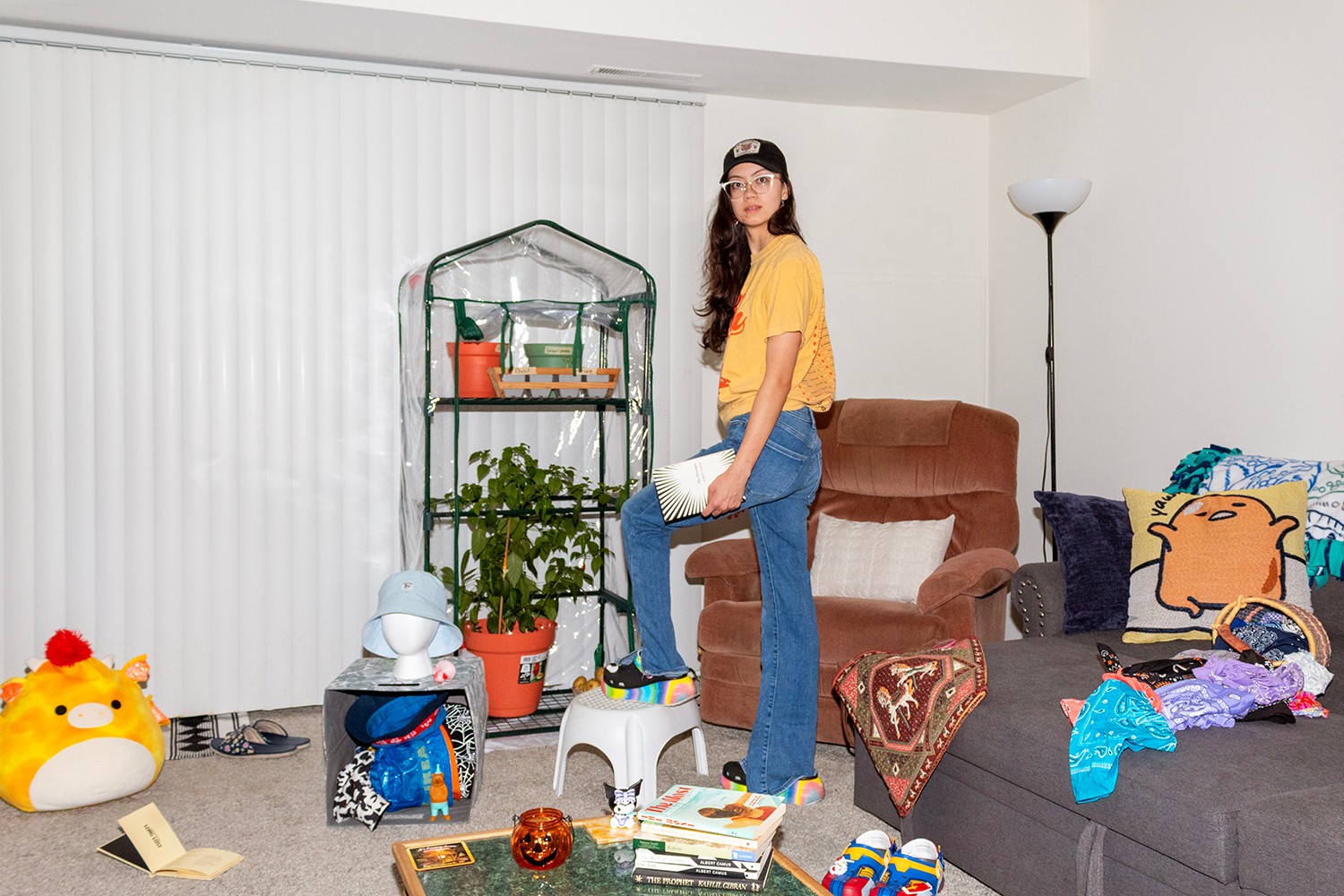 Young woman standing on a sofa in a living room with plants, toys, and colorful cushions, facing camera.