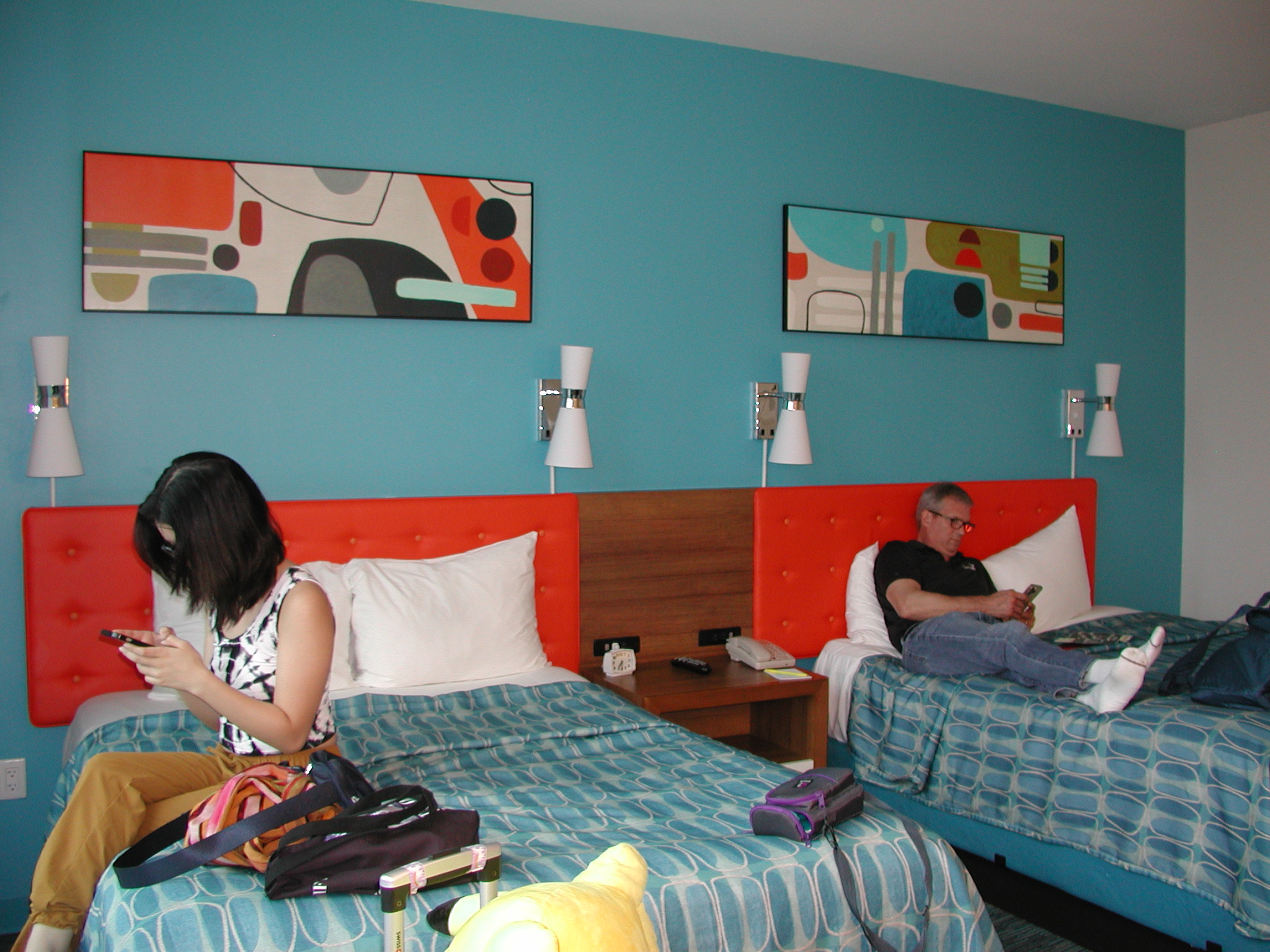 Two beds with red headboards and blue bedding in a colorful room, one person sitting on each bed, wall art above, nightstand between beds.