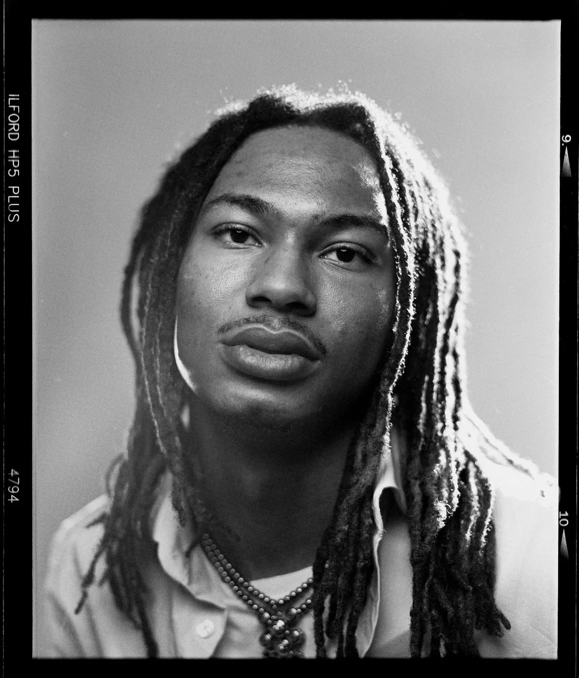 Black and white portrait of a person with long dreadlocks, wearing a collared shirt and necklaces, looking at the camera.