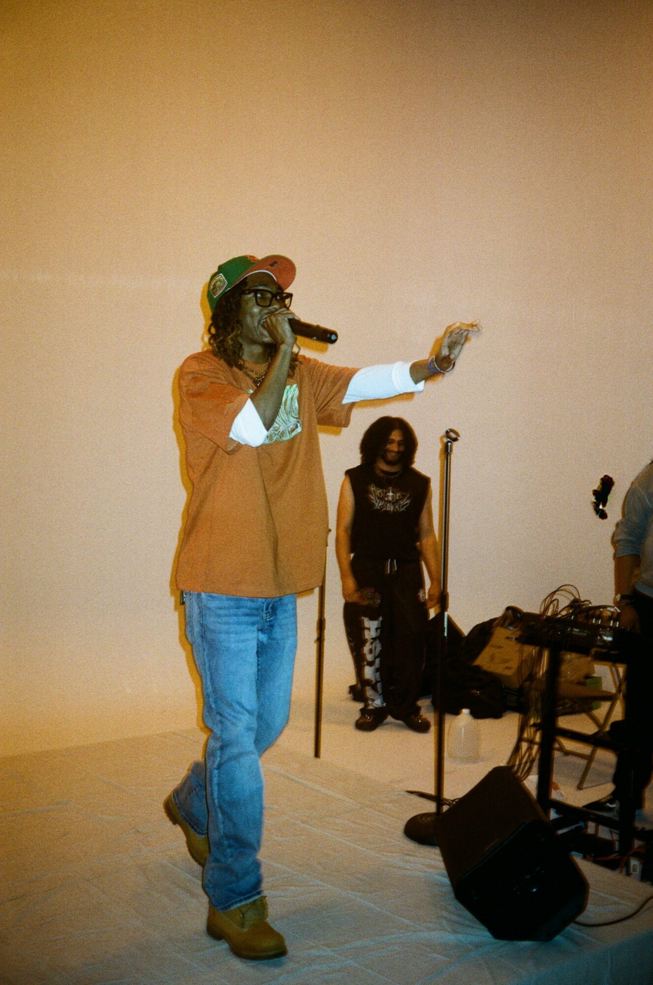 Person wearing glasses, orange jacket, and cap, holding a microphone and gesturing with their right hand, on stage.
