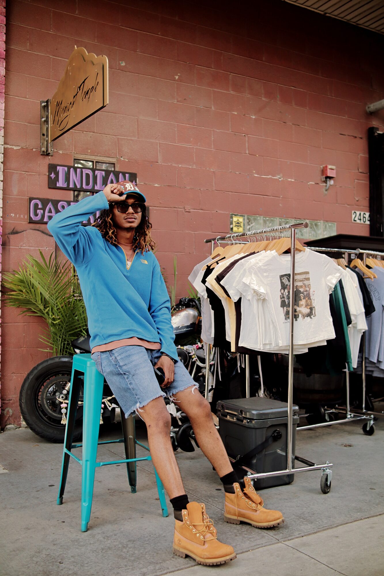 Person with sunglasses and curly hair sitting on a turquoise stool, wearing a blue hoodie, denim shorts, and tan boots, in front of a brick wall with clothing racks.