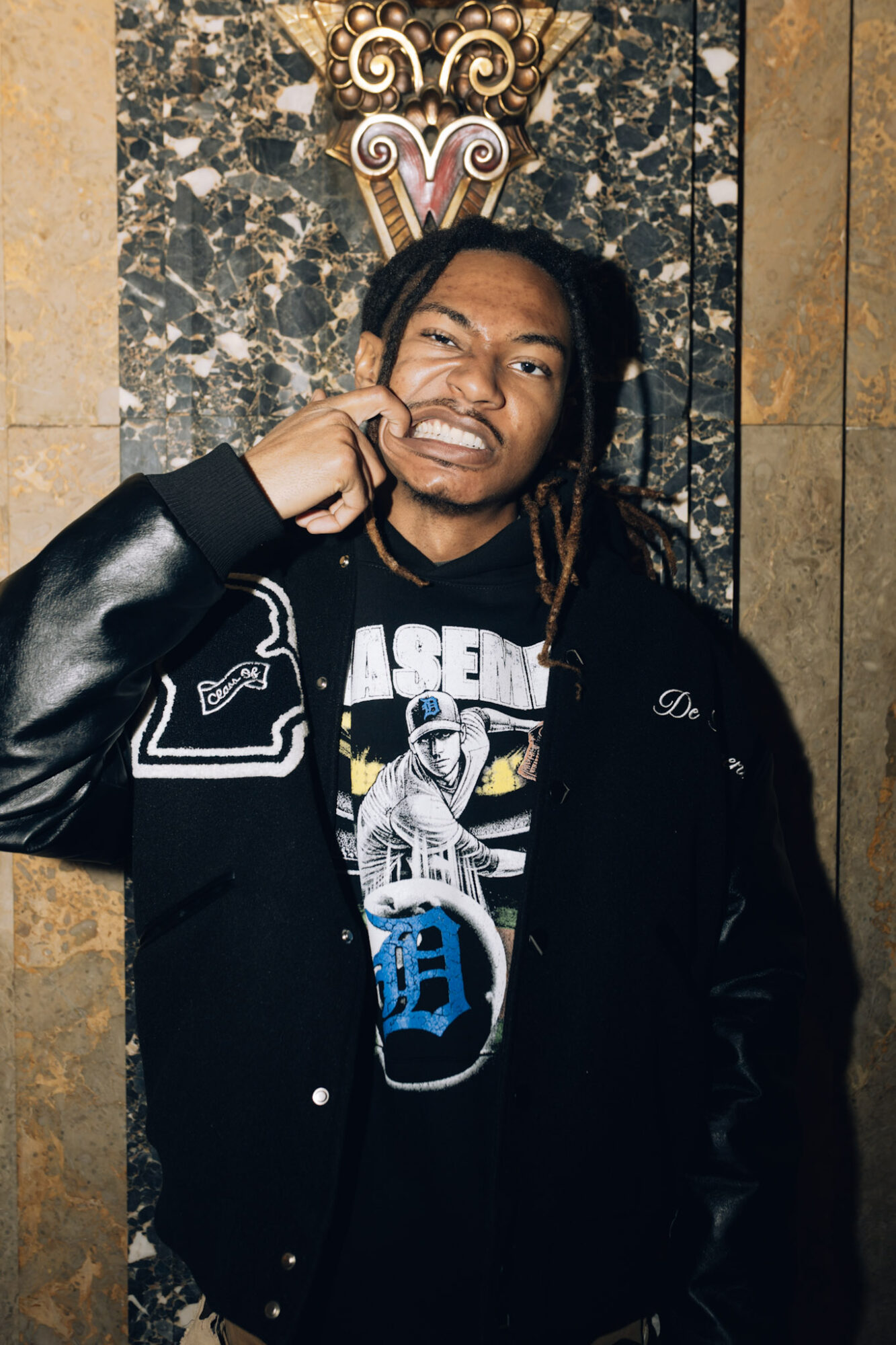 Young man with dreadlocks wearing a black jacket and graphic t-shirt, standing against a decorative wall background.