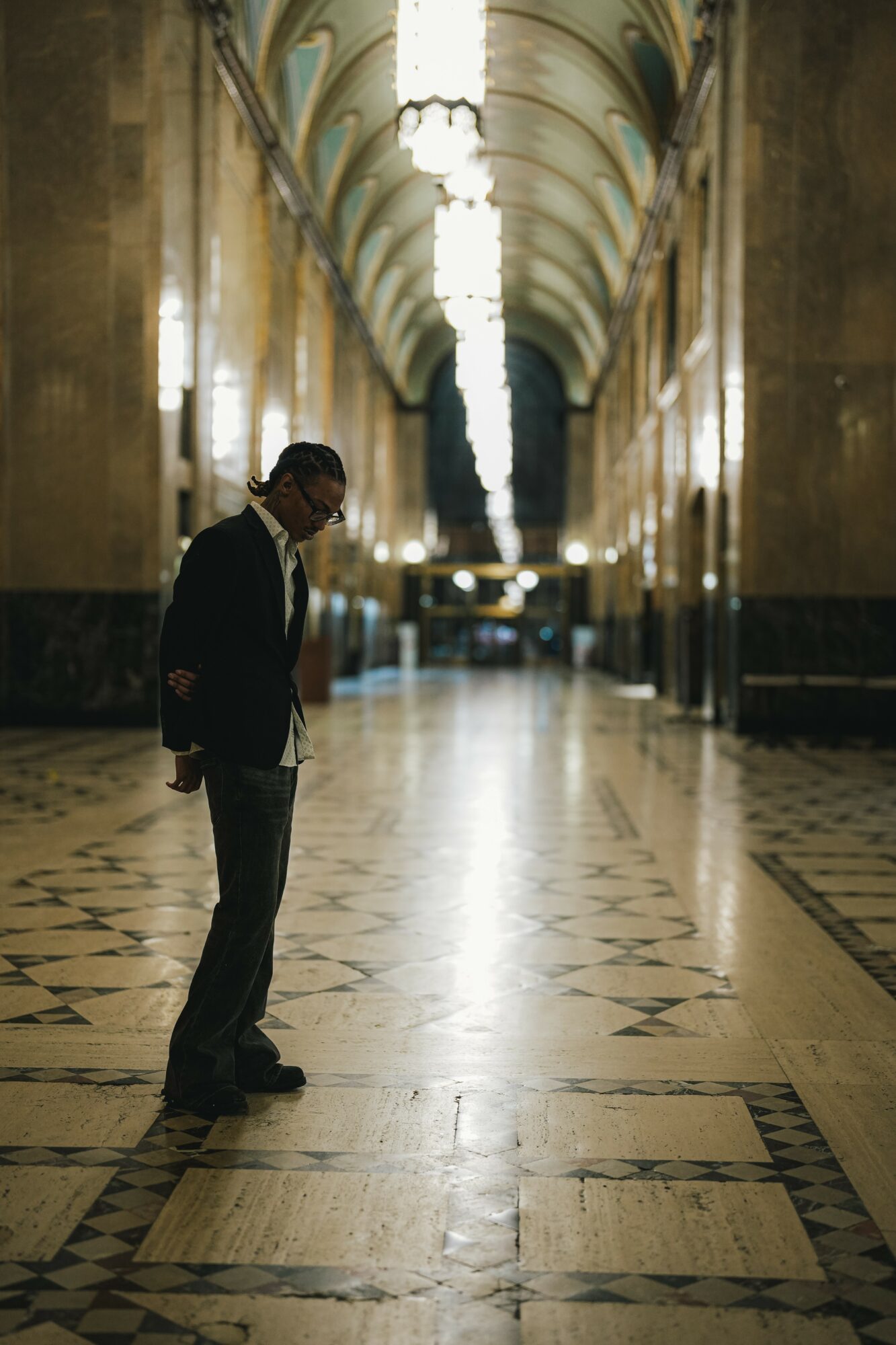 Man standing in a grand, ornate hallway with high arched ceilings and a patterned floor, illuminated by a large chandelier.