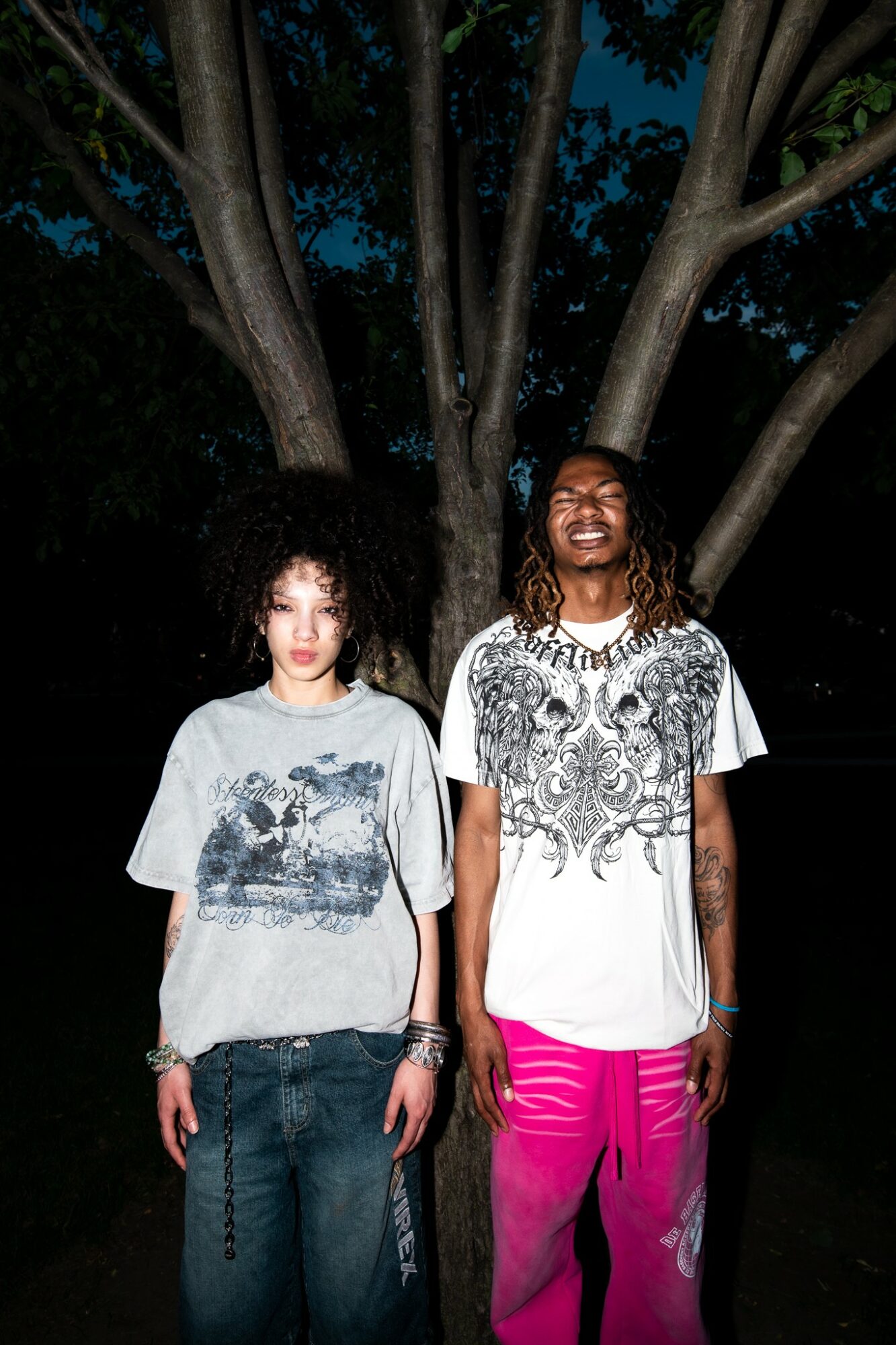 Two women stand outdoors at night in front of a large tree, smiling, with dark sky and foliage behind them.