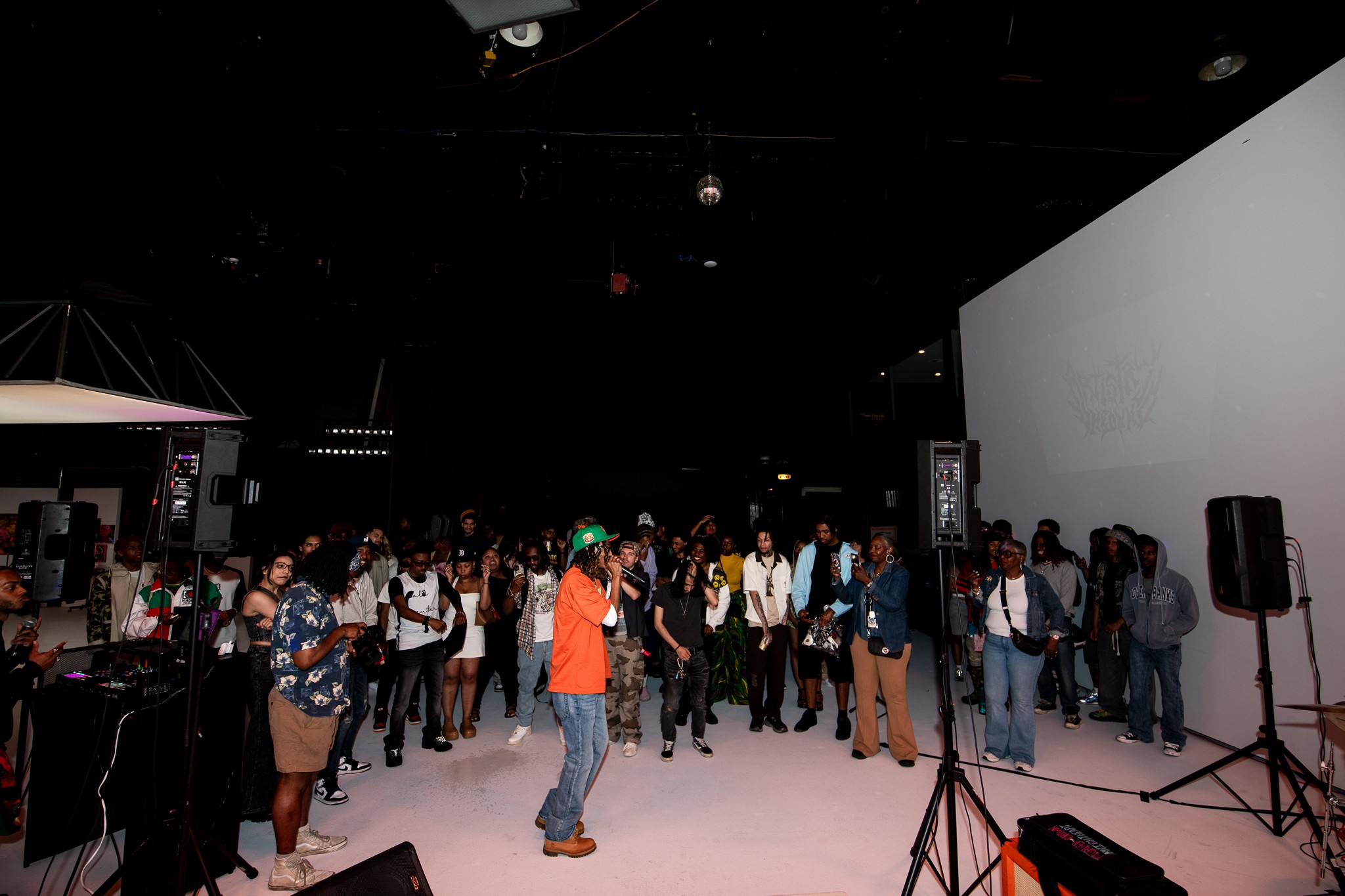 People gather in a dark indoor space with a large white screen and speakers, some standing and talking, others walking.