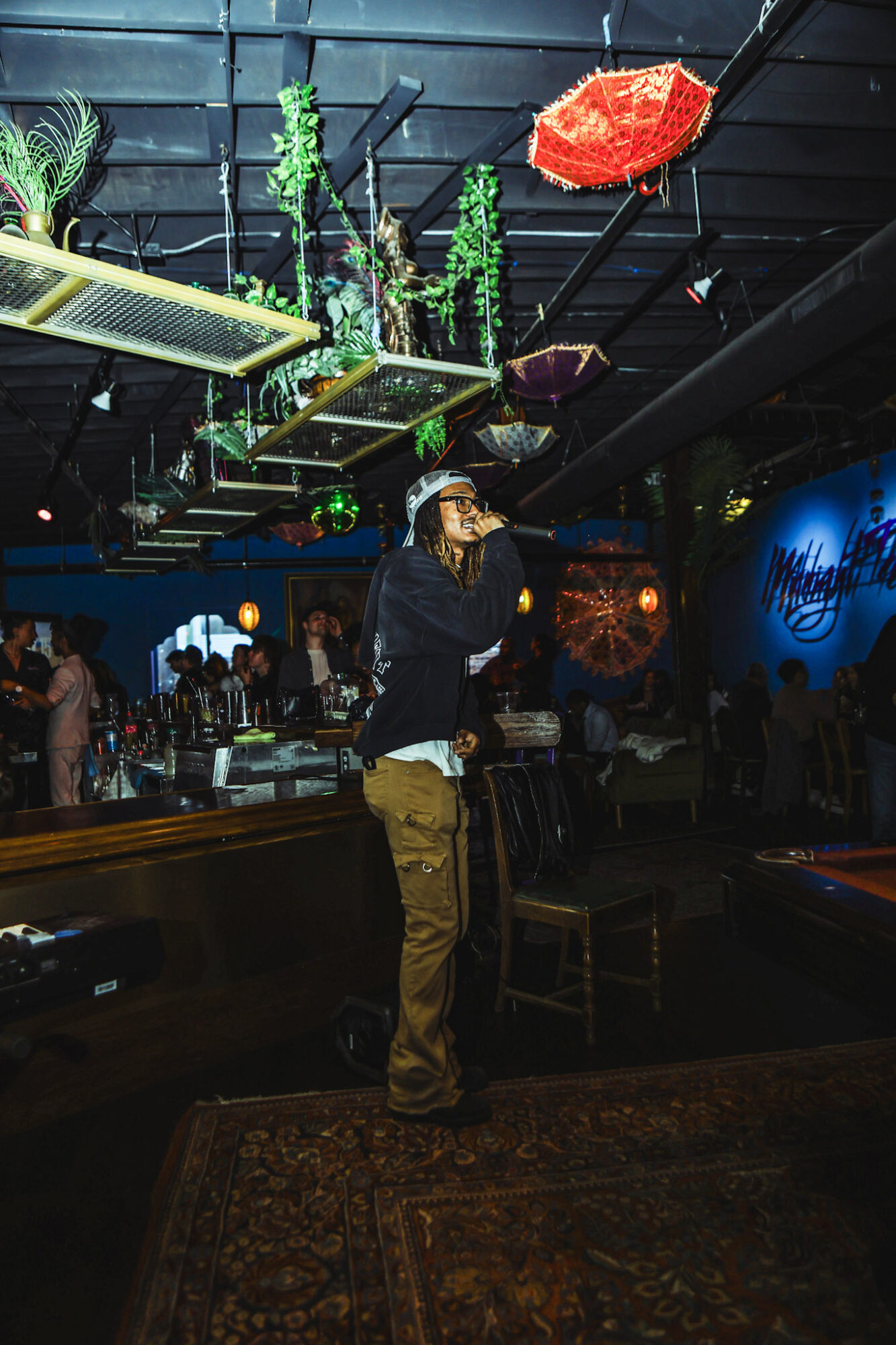 Person standing in a dimly lit bar or club with colorful hanging decorations and a patterned rug.