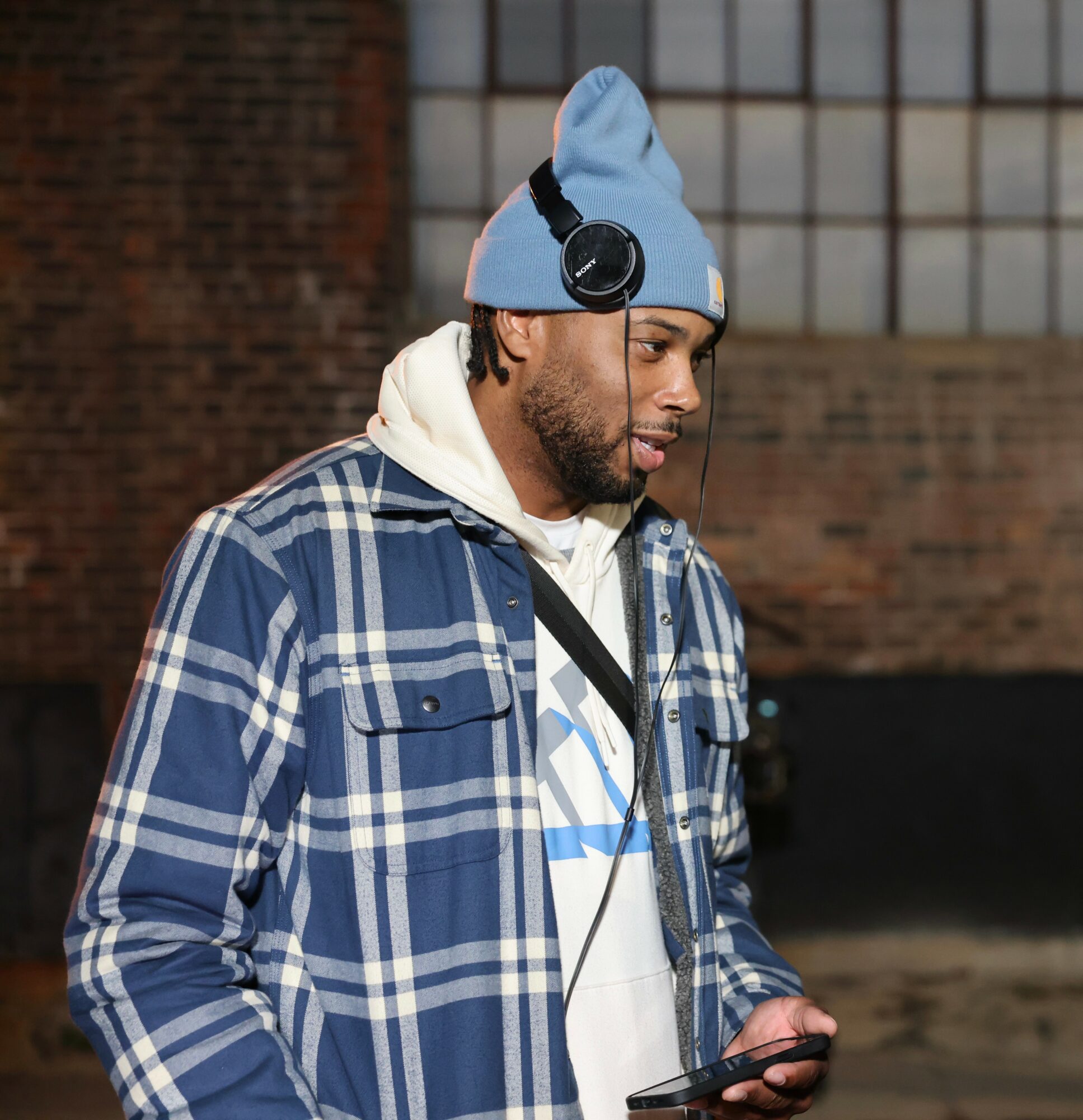 Man wearing a blue beanie, headphones, and a plaid jacket, looking at a phone outdoors.