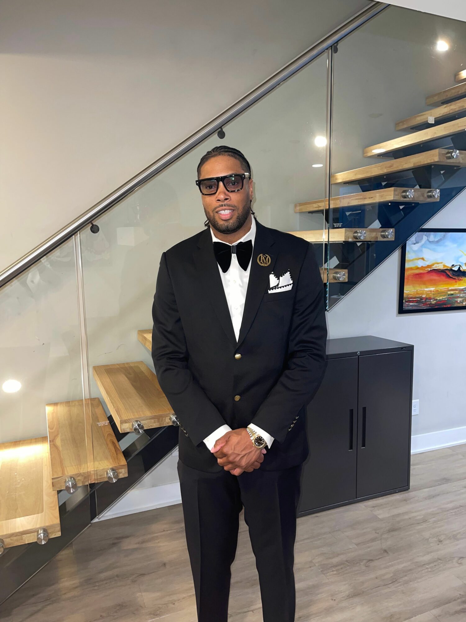 Man in black tuxedo with bow tie and sunglasses standing indoors, smiling, with a staircase and TV in background.