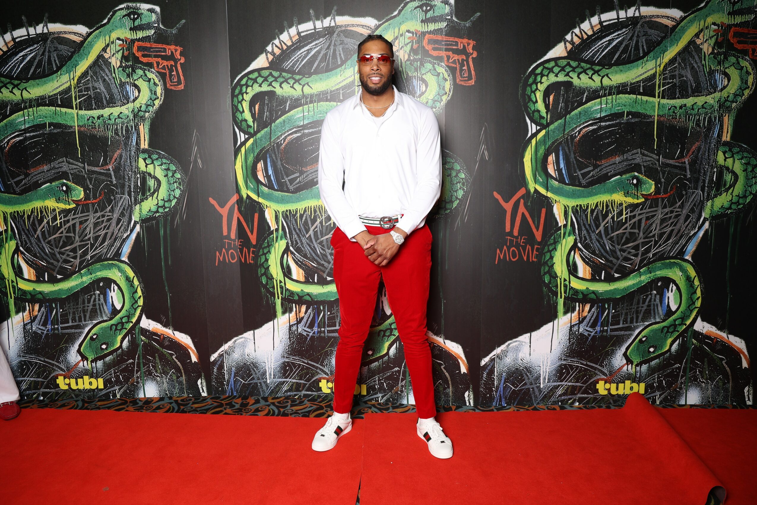 Man in white shirt and red pants standing on red carpet in front of a snake-themed mural.