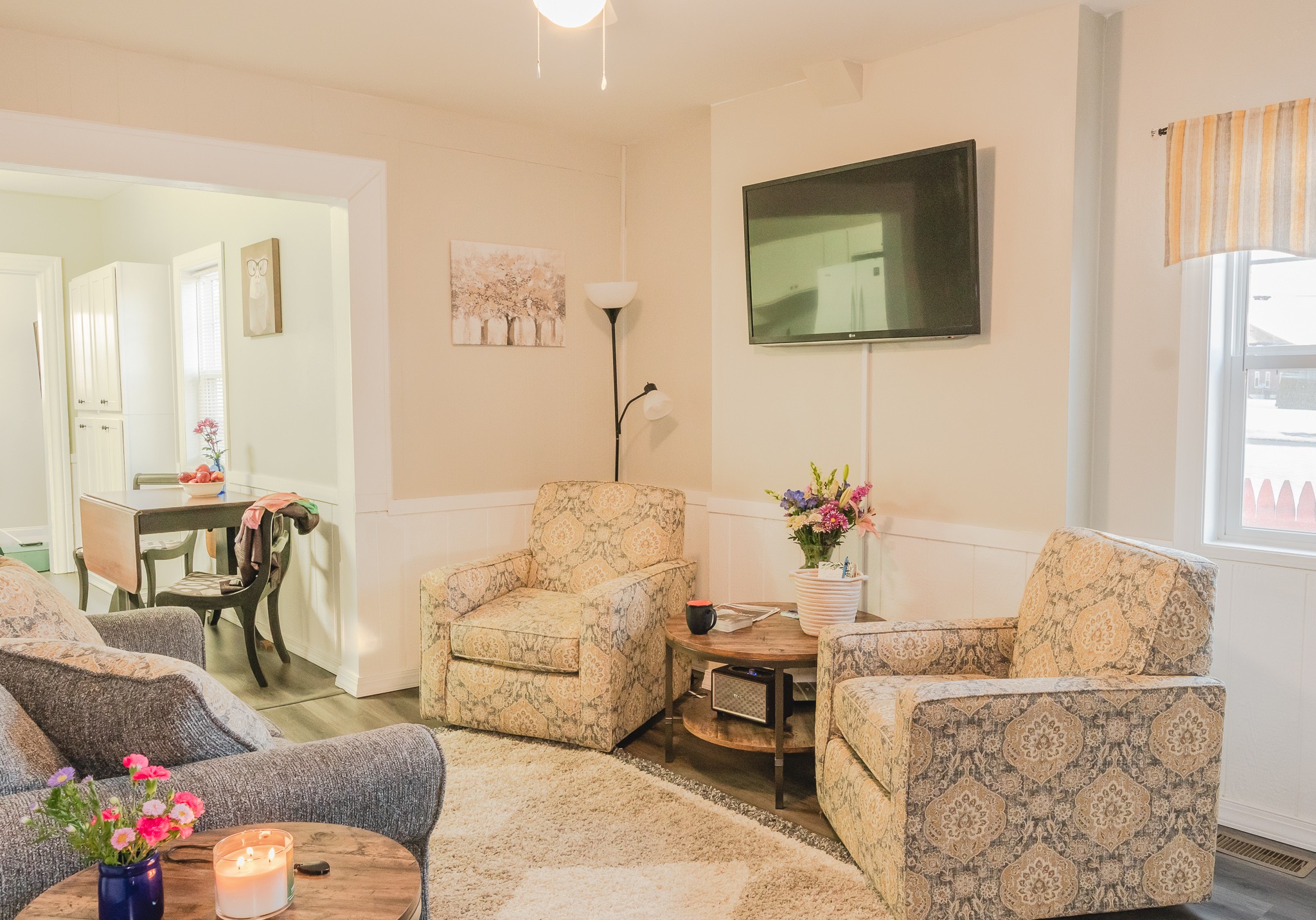 Living room with two patterned armchairs, a side table with flowers, a wall-mounted TV, and a window with curtains.