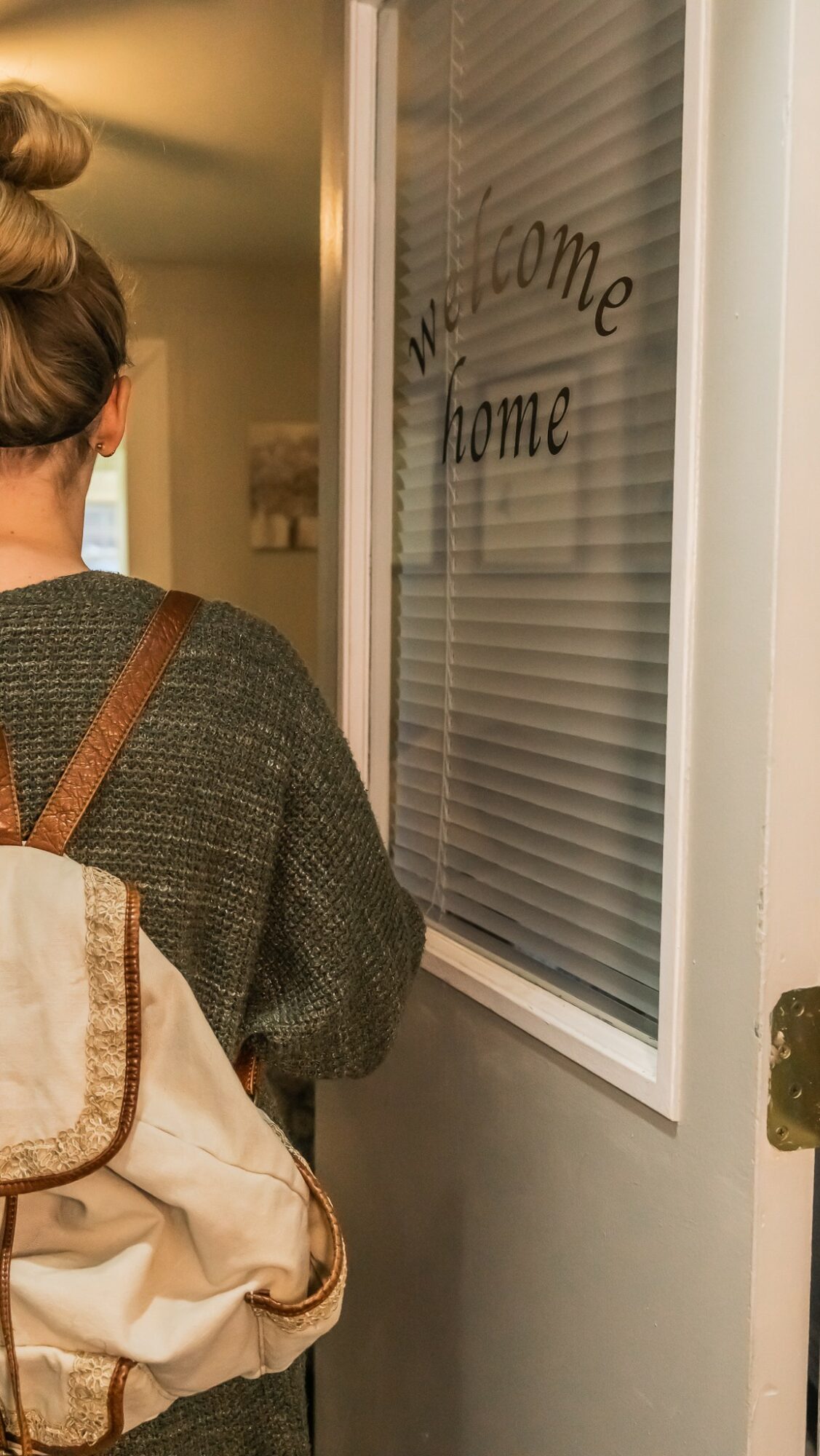 Person with brown hair in a bun, wearing a dark sweater, standing near a door with a window that says 'Welcome Home'.