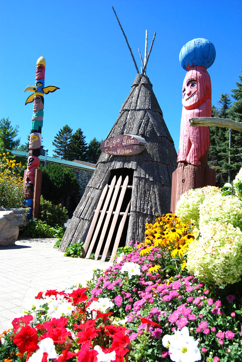 Colorful totem poles and a thatched hut surrounded by vibrant flowers and greenery under a clear blue sky.